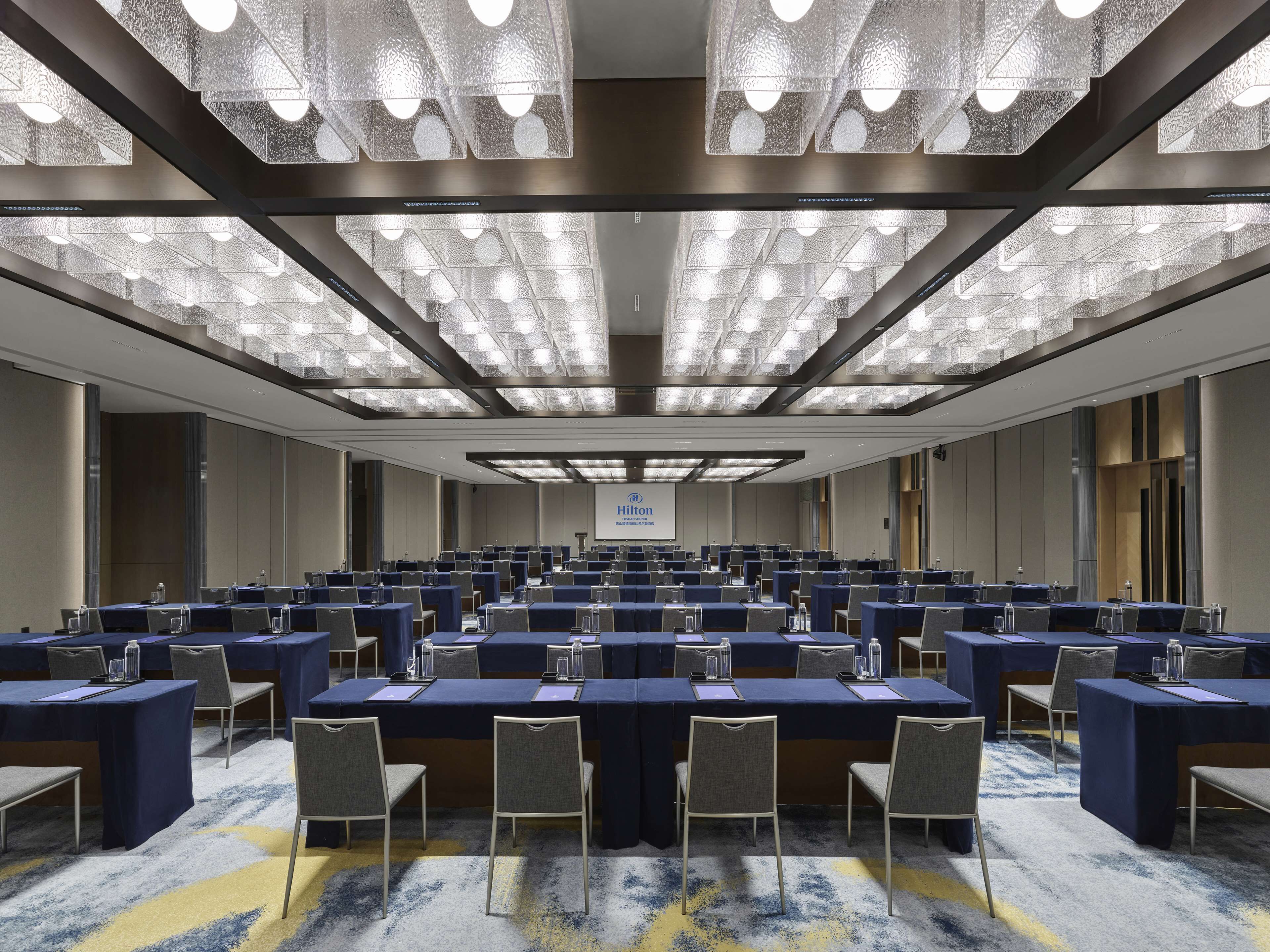 A large conference room with multiple tables and chairs, a carpeted floor, and a ceiling with patterned lighting fixtures.