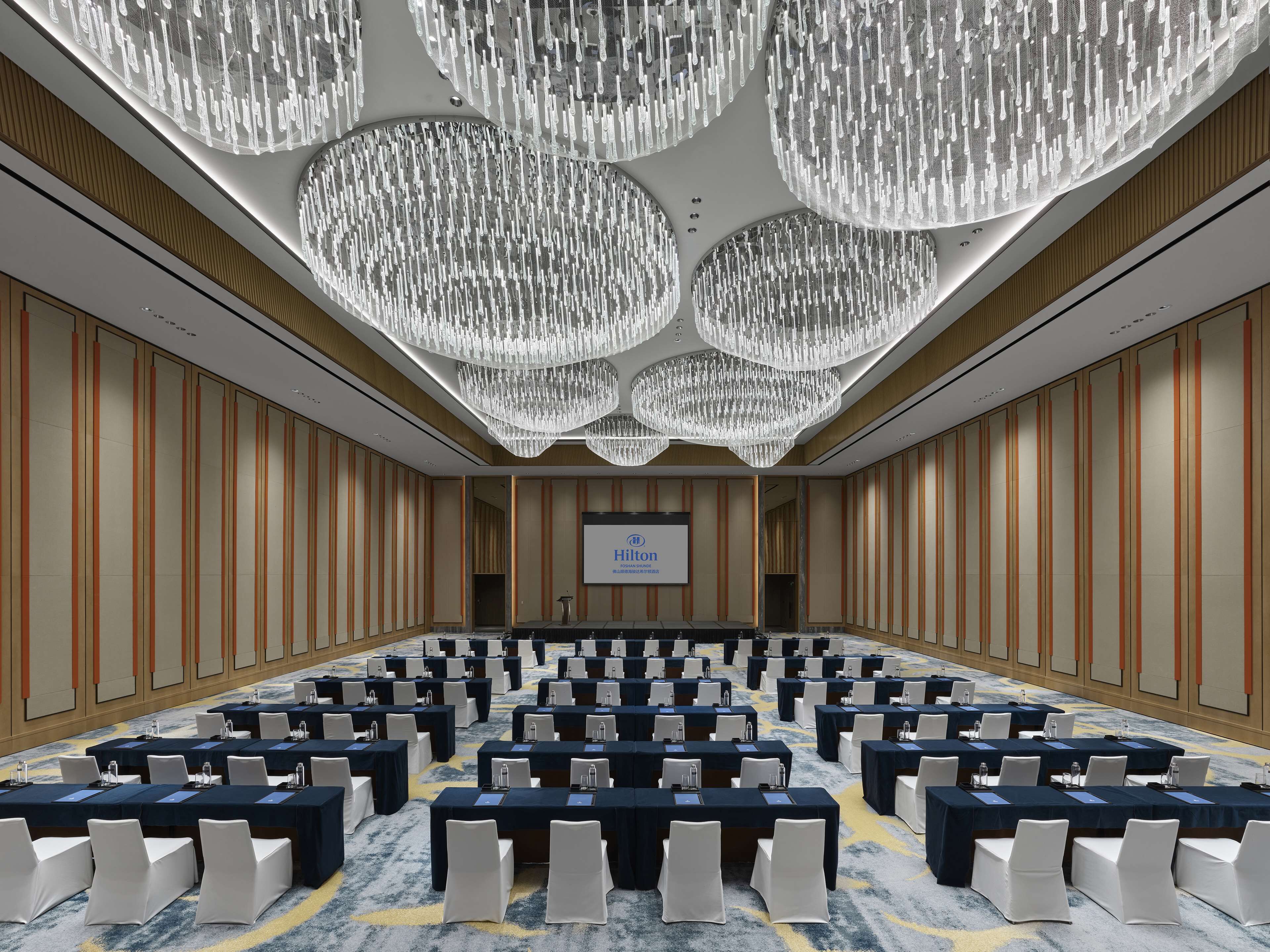 A large conference room with rows of tables and chairs, a large screen, and a chandelier.