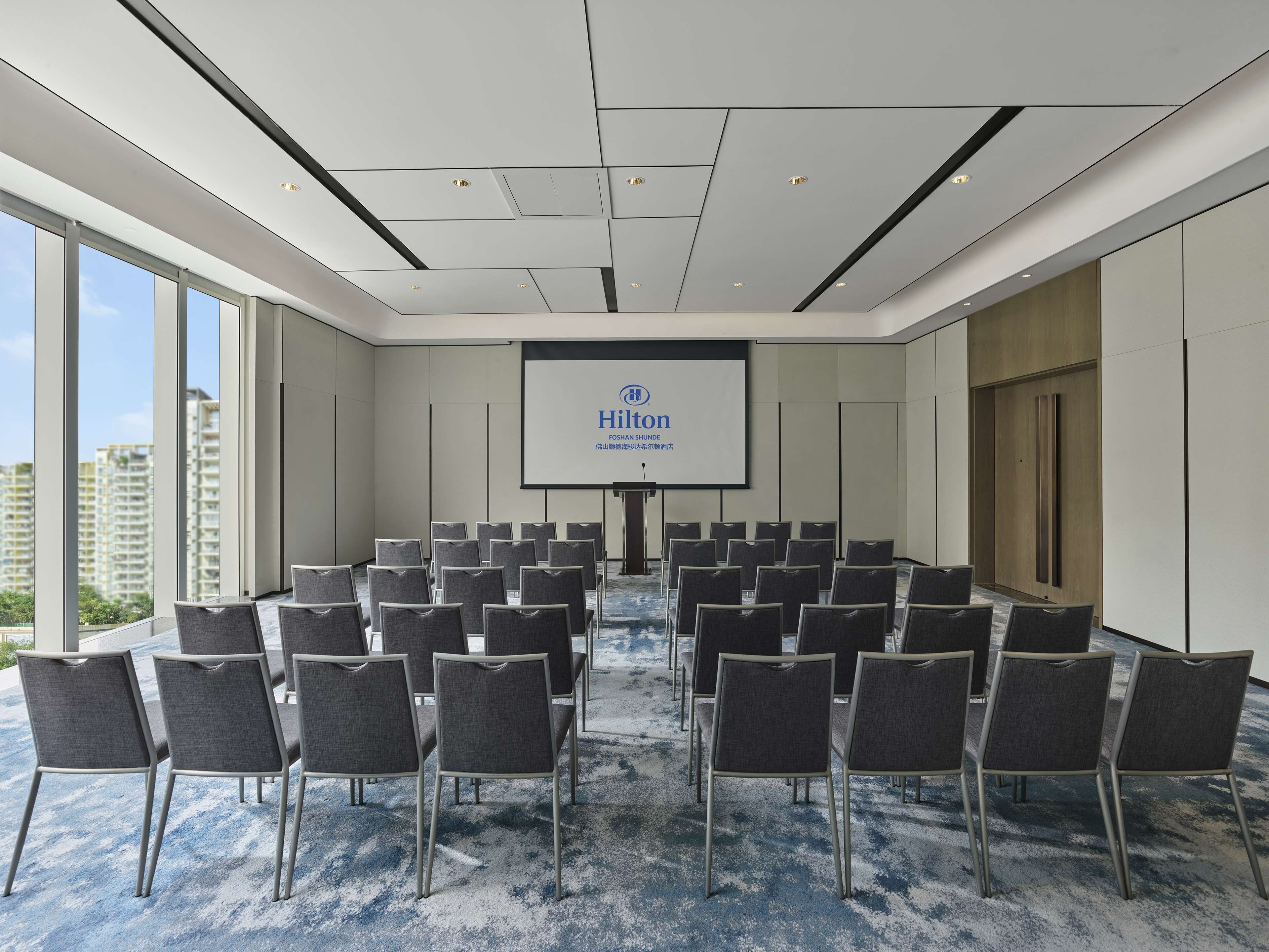 A conference room with rows of chairs, a large screen displaying the Hilton logo, and a city view through large windows.