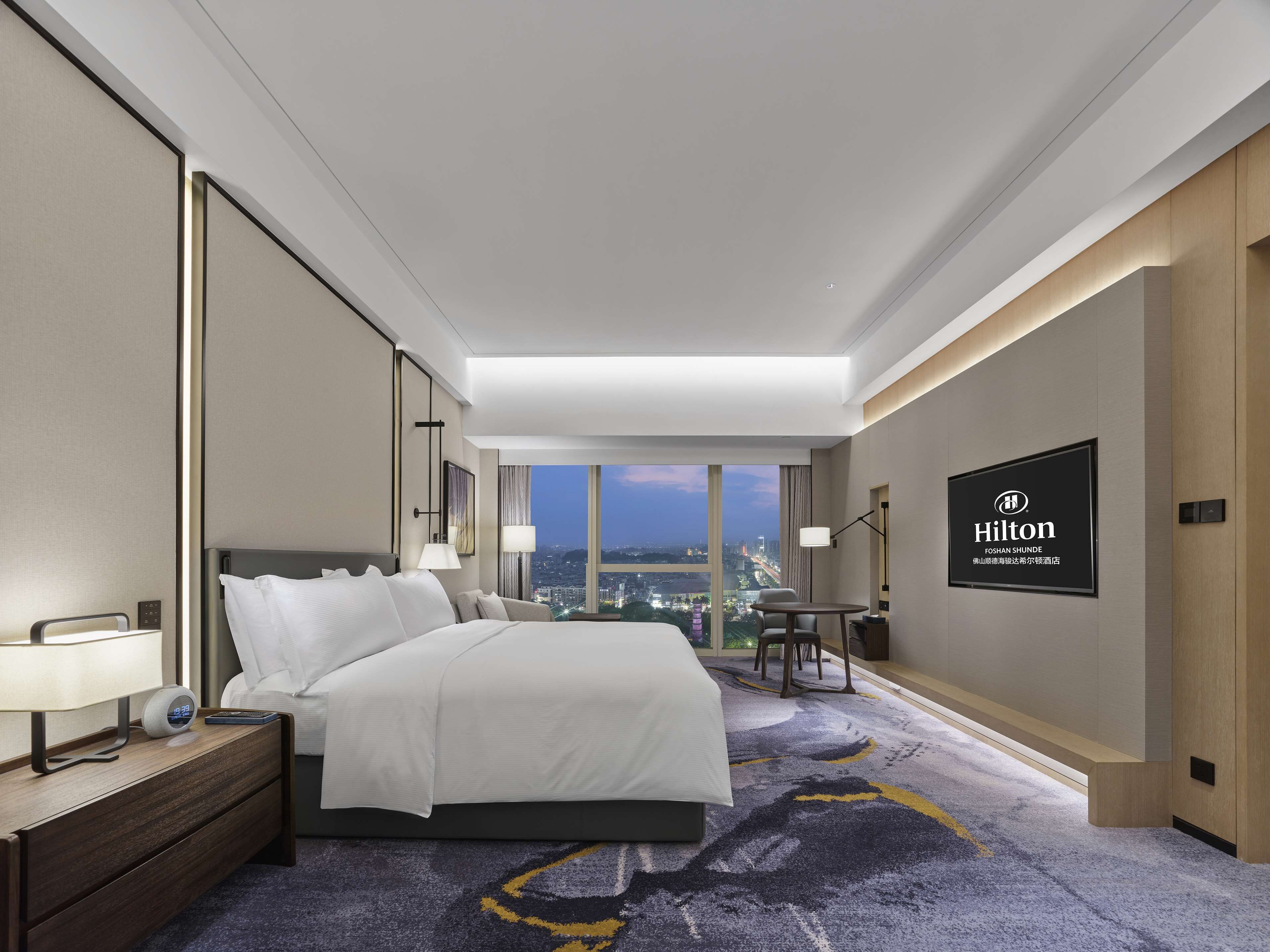 Une chambre d’hôtel moderne, dotée d’un grand lit, de tables de chevet, d’un bureau et offrant une vue sur la ville au crépuscule.