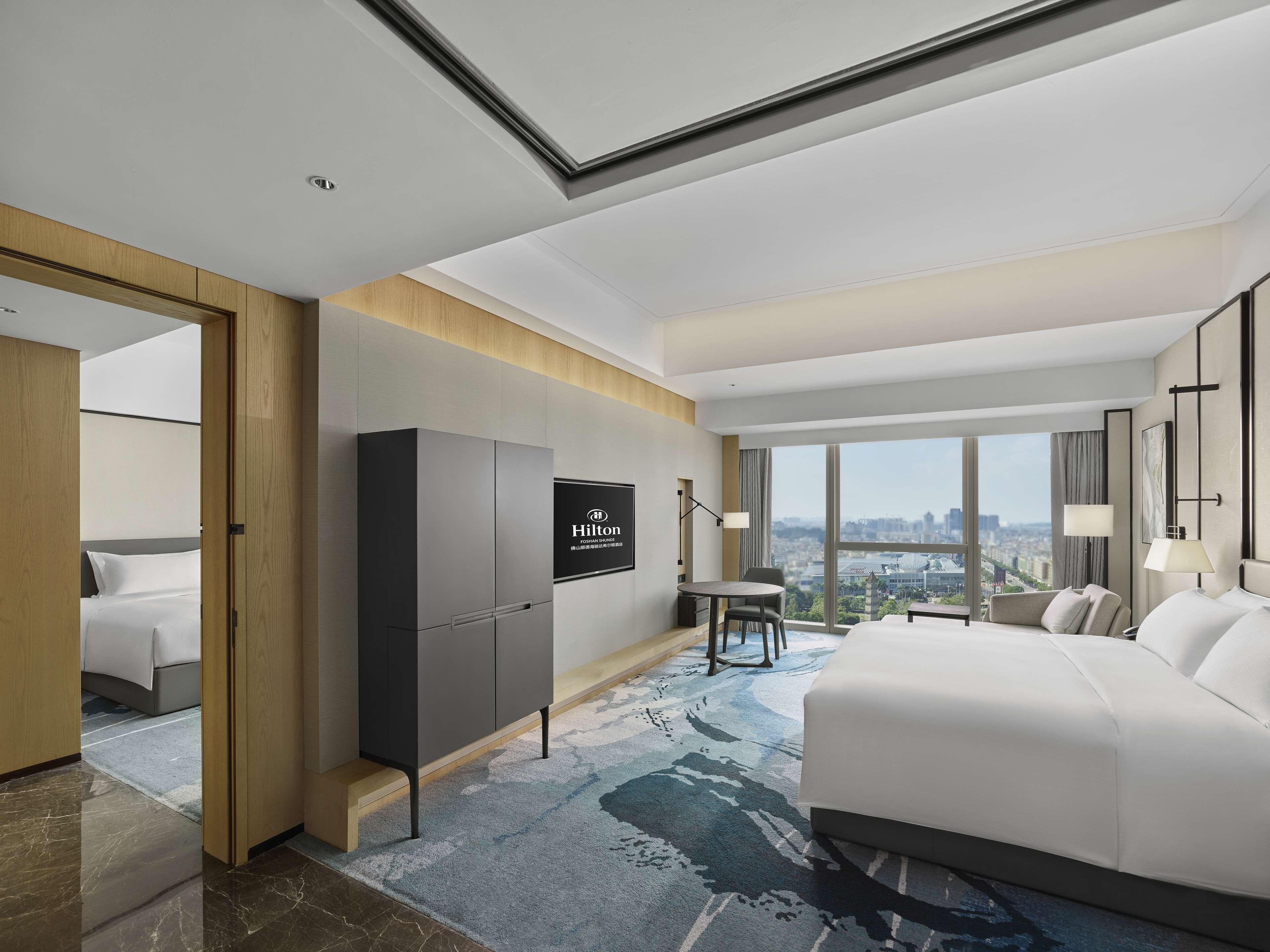 A modern hotel room with a large bed, a seating area with a table and chairs, a minibar, and a city view through large windows.