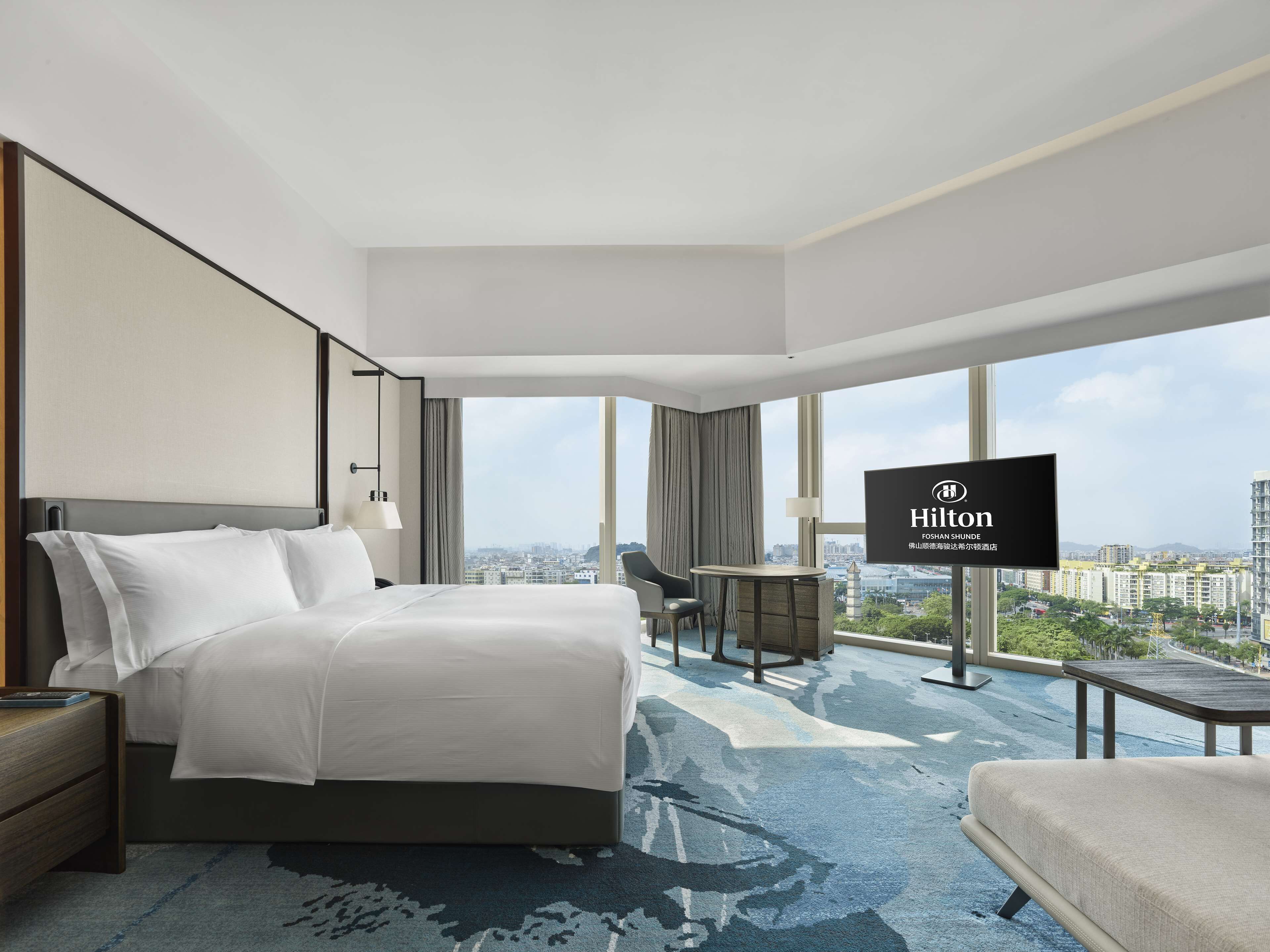 A modern hotel room with a large bed, a desk, a chair, and a TV displaying the Hilton logo.