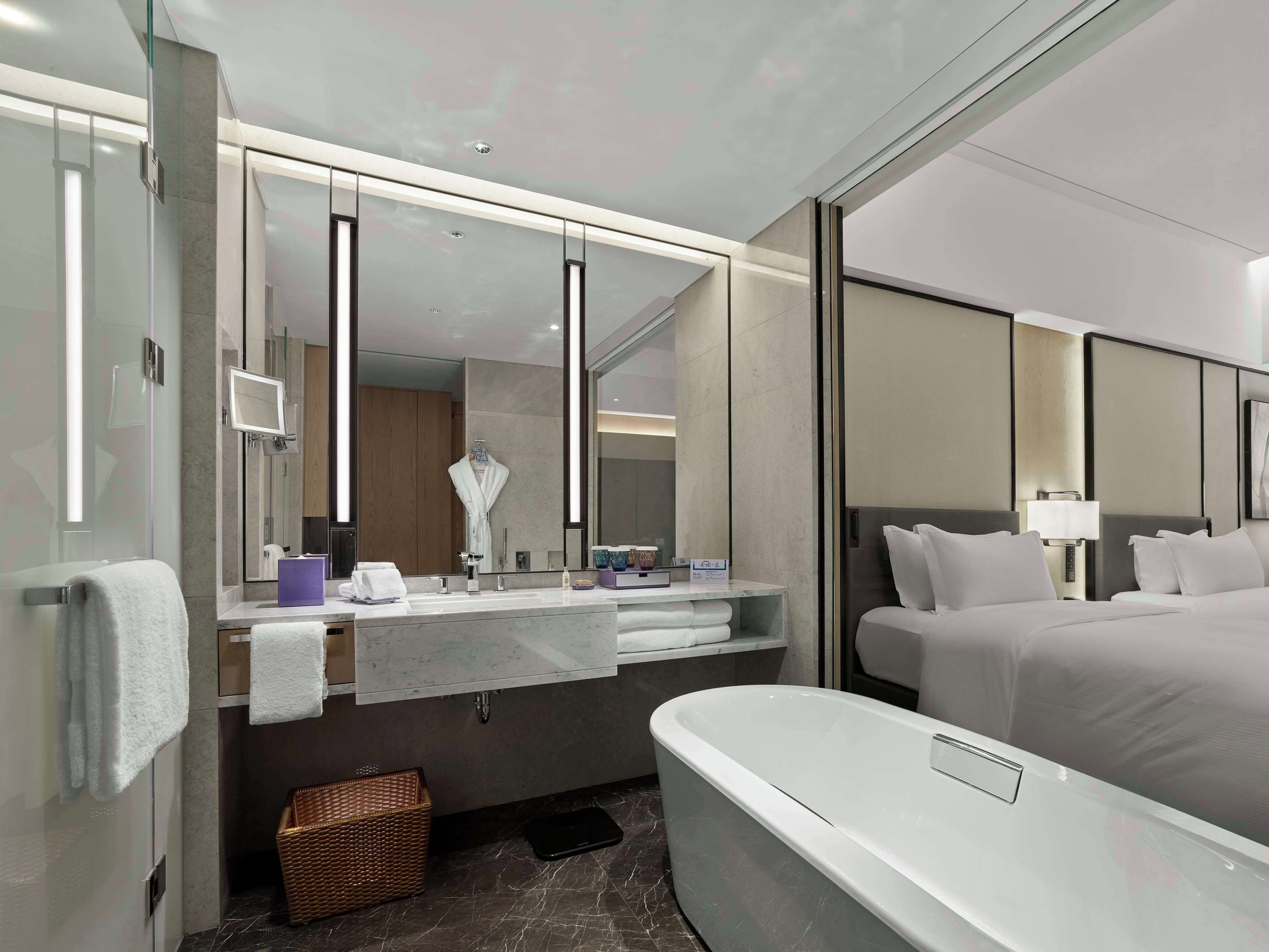 A modern hotel room with a large mirror, a bathtub, a bed with white linens, and a vanity with a mirror and a sink.