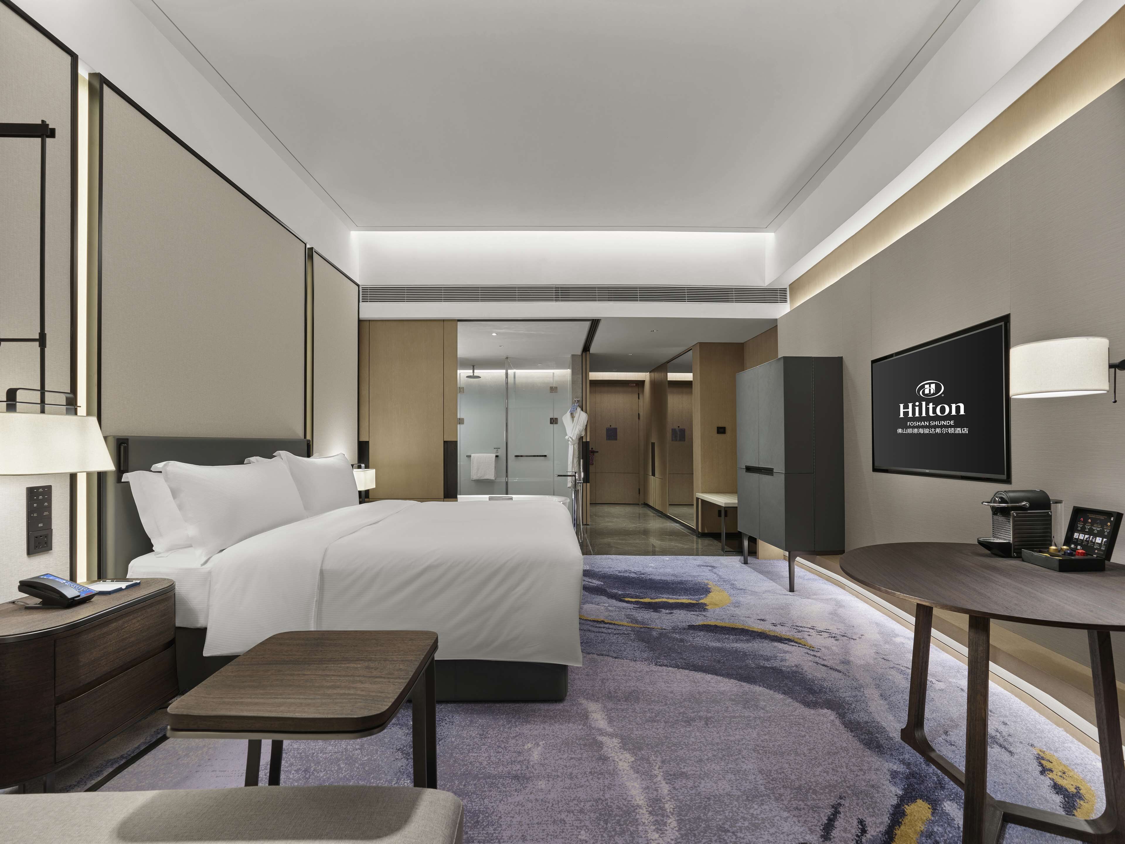 A modern hotel room with a large bed, a desk, a TV, and a bathroom visible in the background.