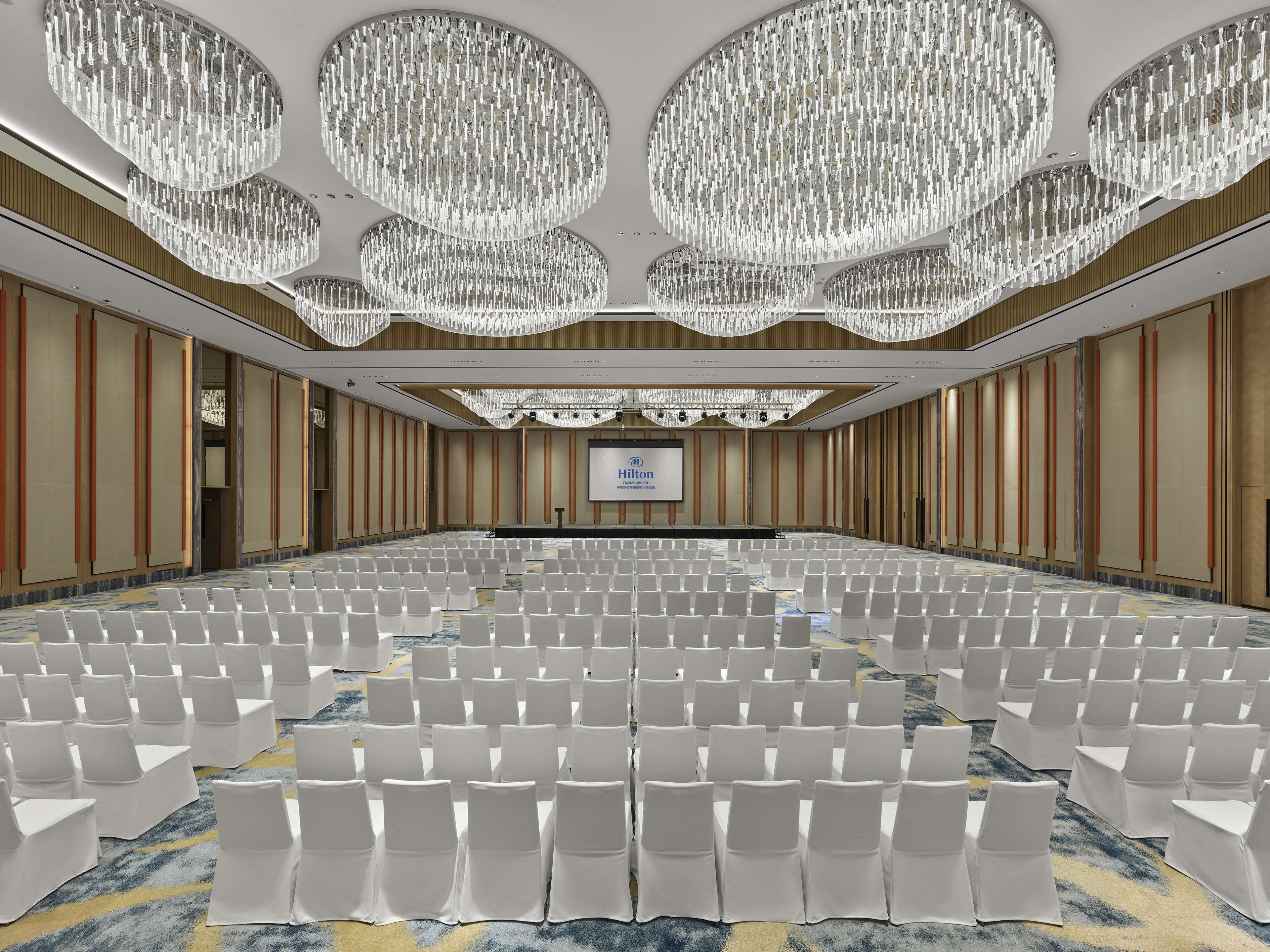 A large conference room with rows of white chairs, a stage with a presentation screen, and ornate chandeliers.