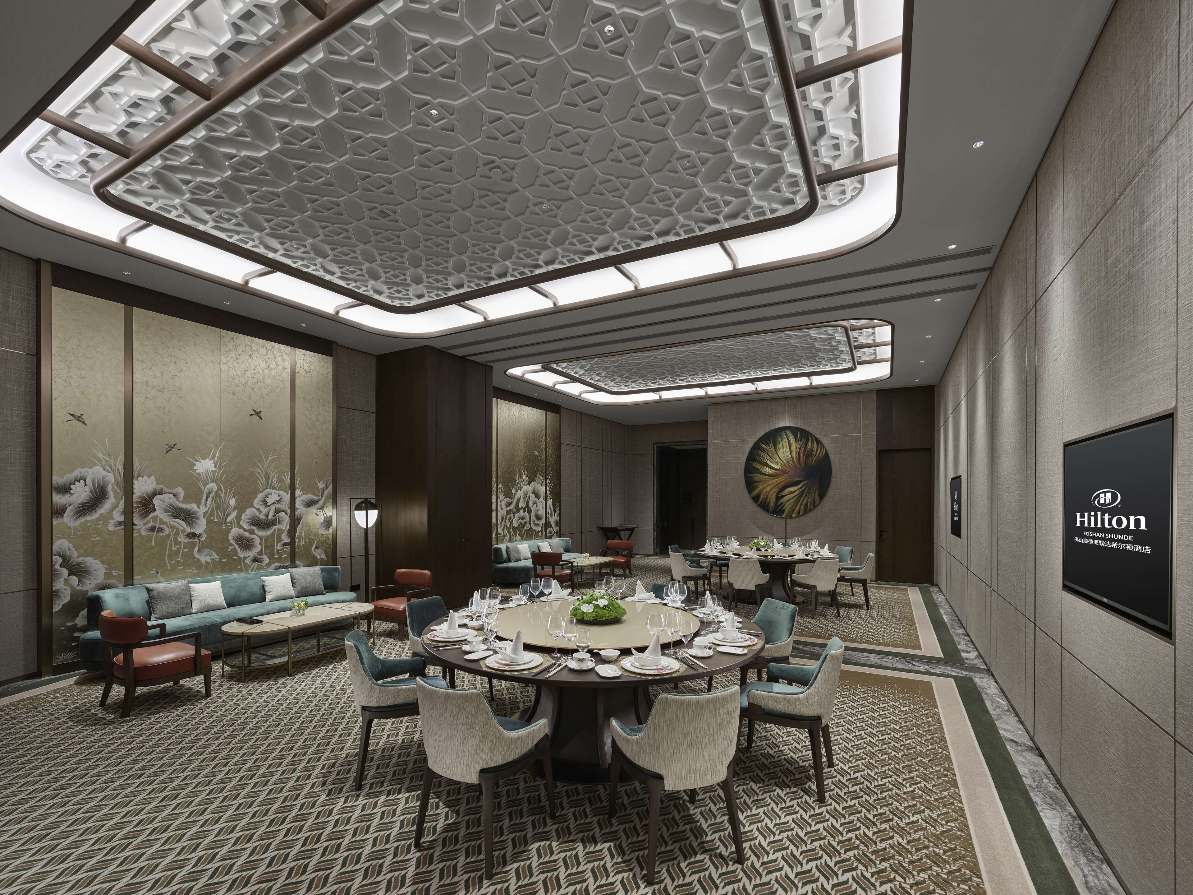 A modern hotel room with a round dining table set for six, a sofa, and a large wall-mounted screen displaying the Hilton logo.