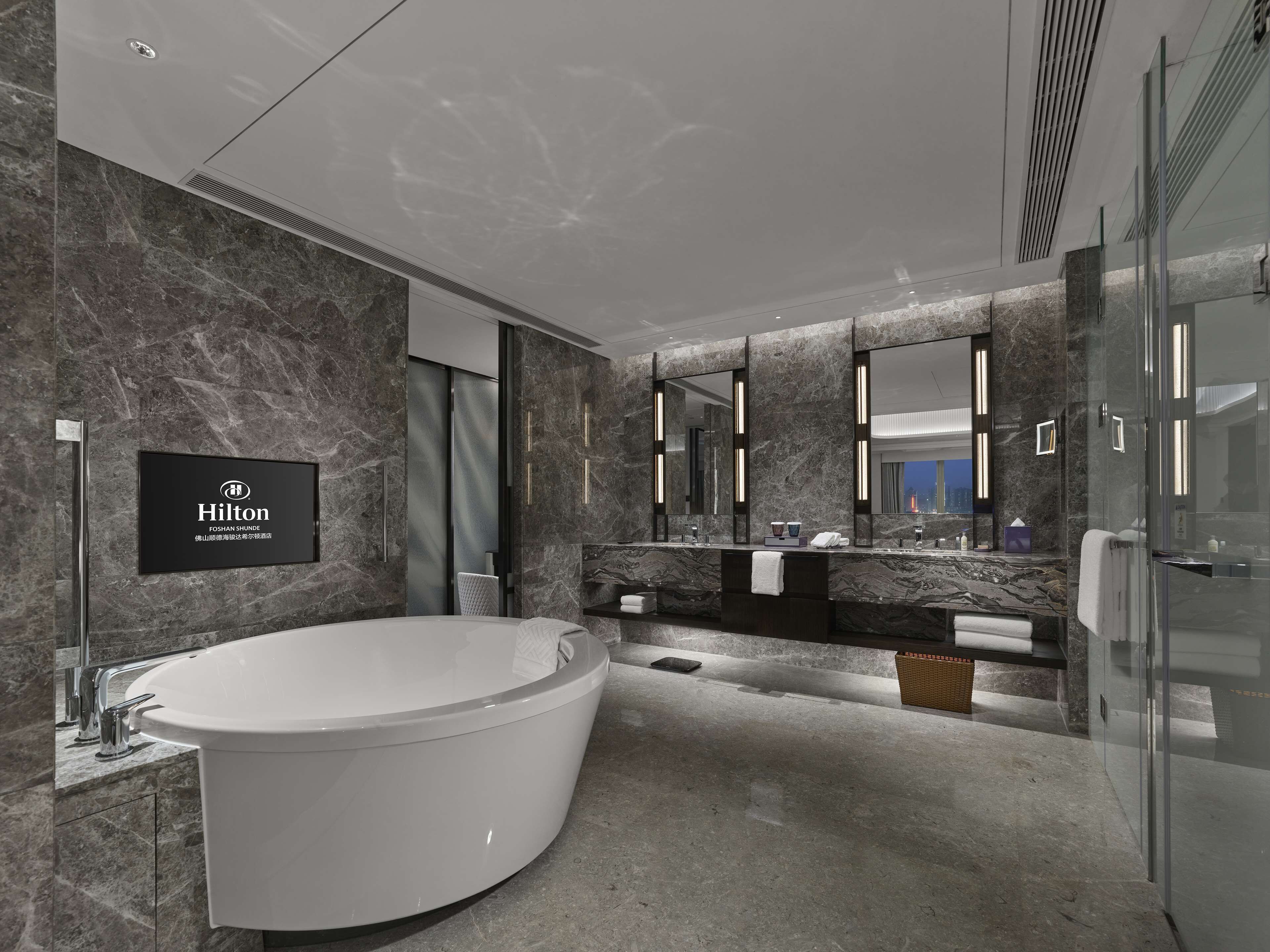 A modern bathroom with a large bathtub, a vanity with a mirror, and a sign indicating it's a Hilton FOSHAN SHUNDE hotel.