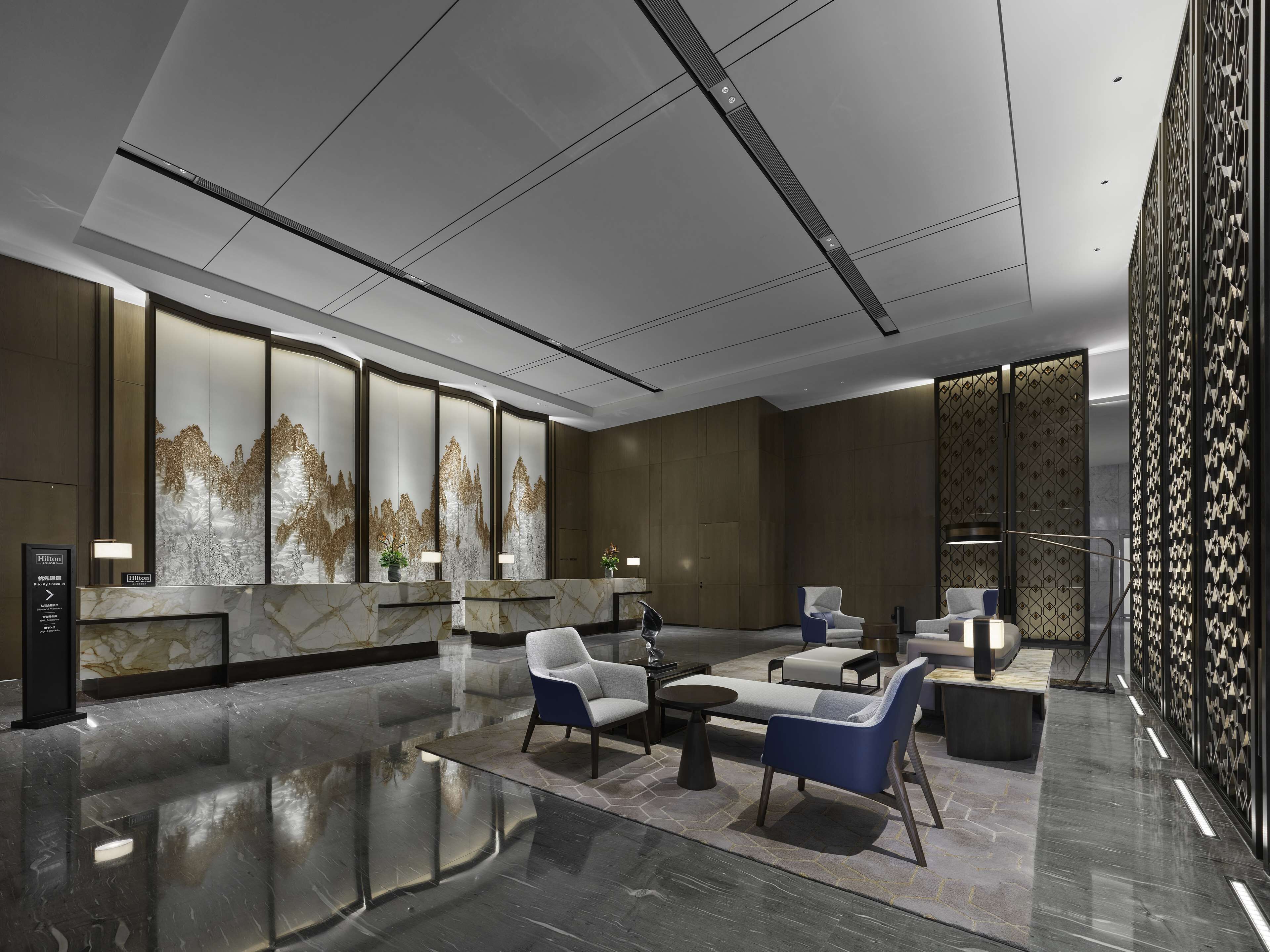A modern lobby with a marble reception desk, seating area with sofas and armchairs, and decorative wall panels.