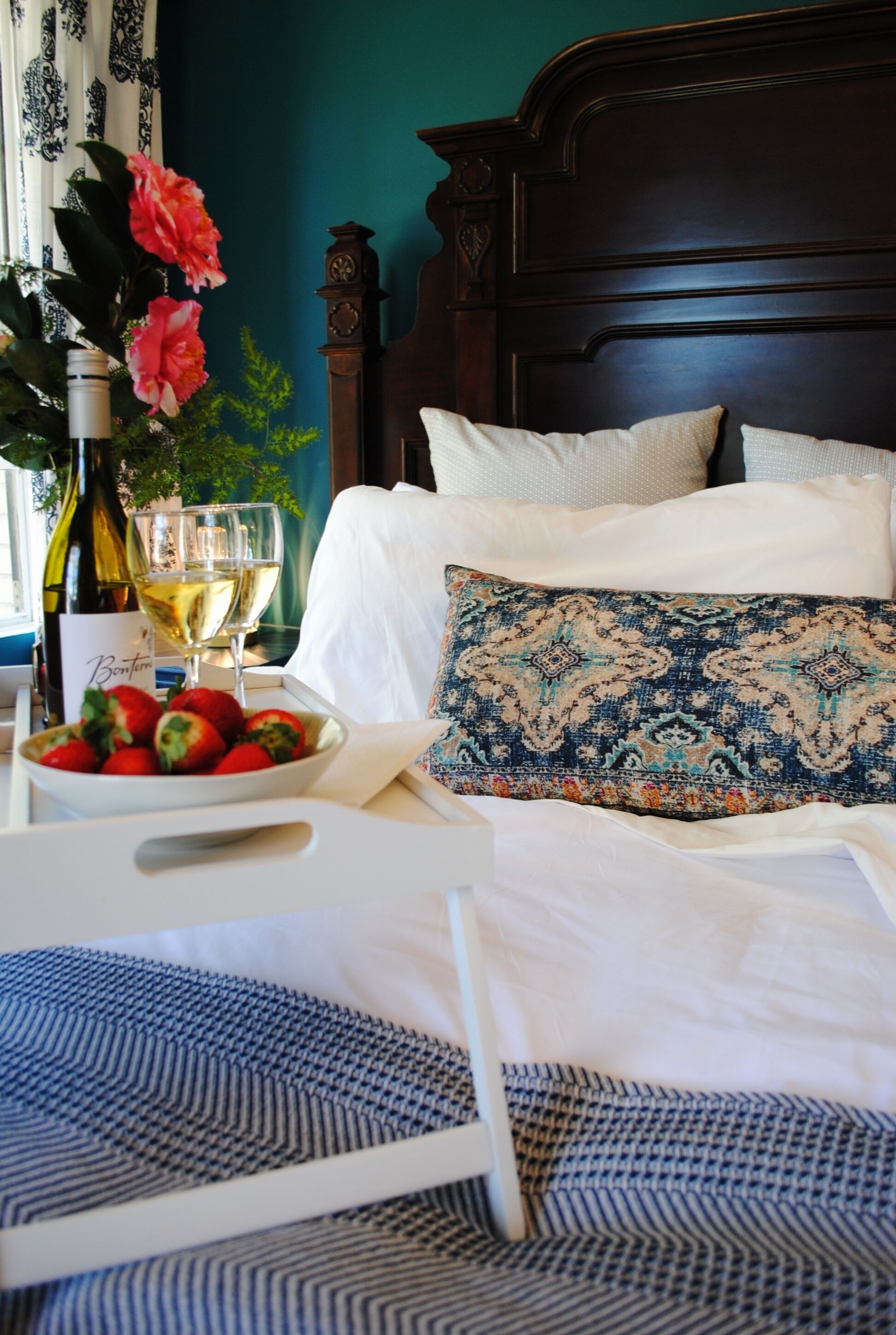 Una cama bien tendida con almohadas blancas y estampadas, un cuenco de fresas, una botella de vino y vasos sobre una mesita de noche.