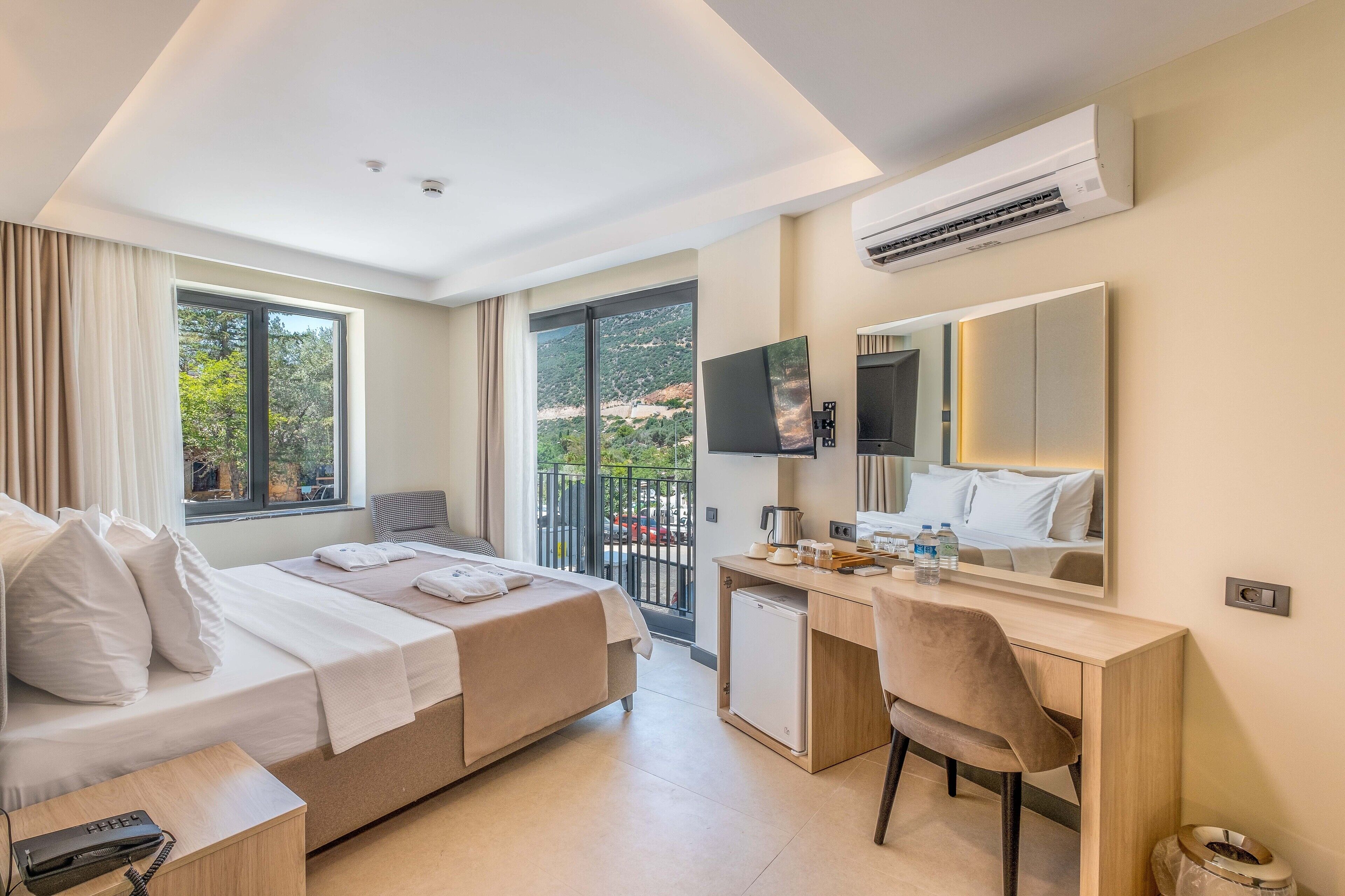 Habitación de hotel con cama, escritorio, silla y un ventanal con vistas a árboles y un balcón.