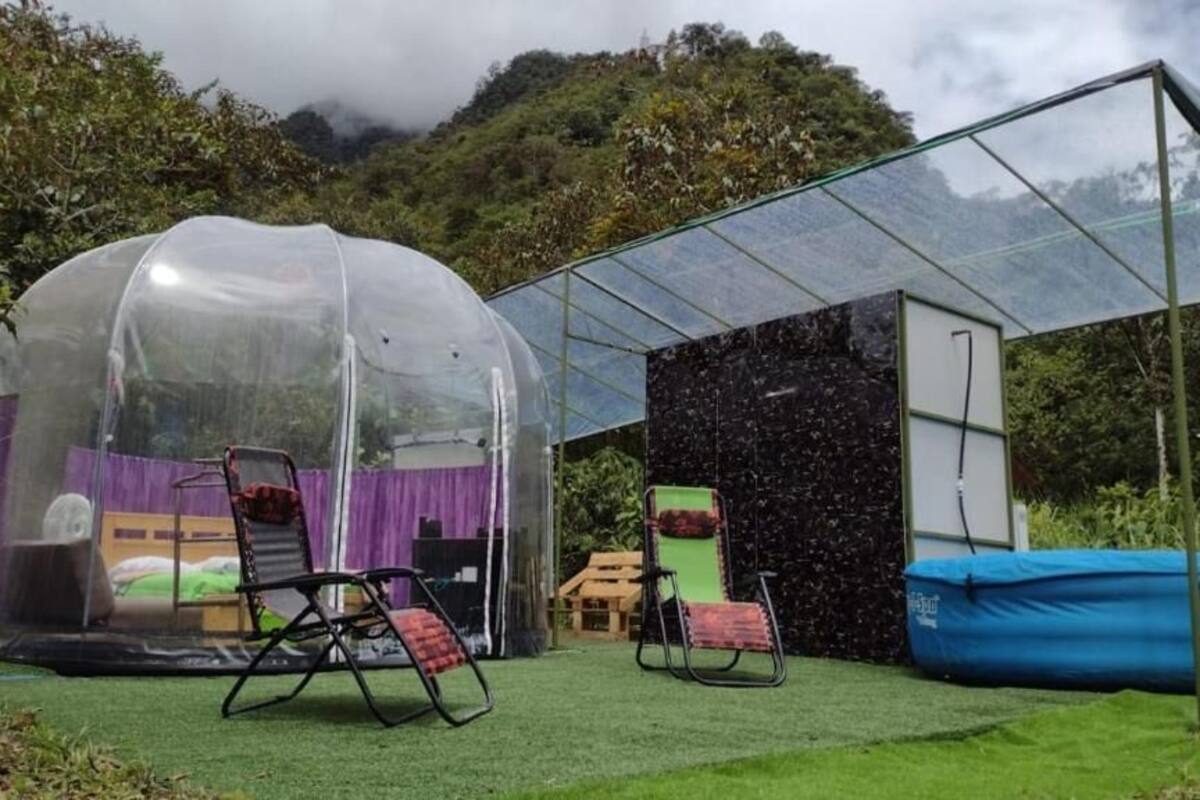 Een transparante tent met een bed en stoelen van binnen, omgeven door een glazen omheining en een zwarte muur.
