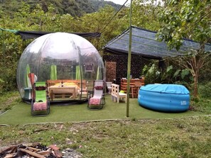 Una estructura abovedada transparente con una mesa y sillas en su interior, una piscina inflable azul y un pequeño edificio con techo inclinado al fondo.
