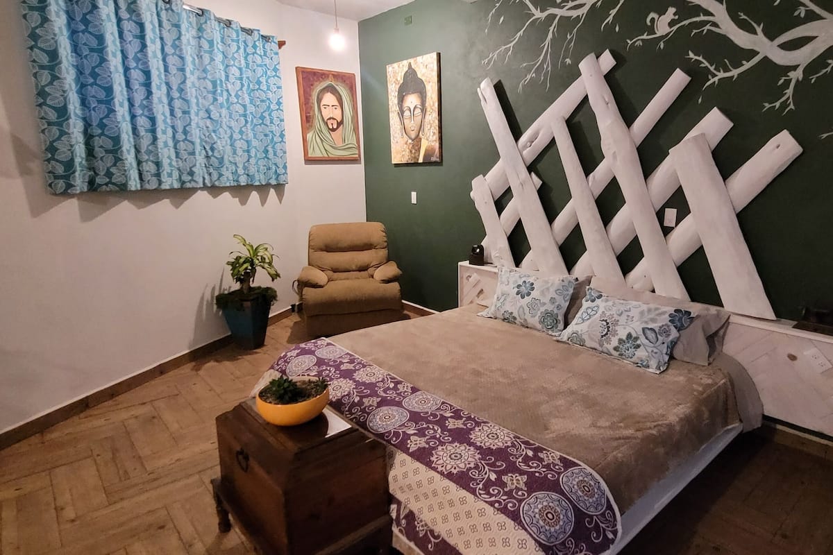 A bedroom with a bed, a chair, a nightstand, a potted plant, and a decorative wall.