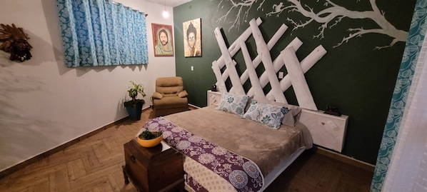 Un dormitorio con cama, una silla, una mesita de noche, una planta en maceta y un adorno en la pared.