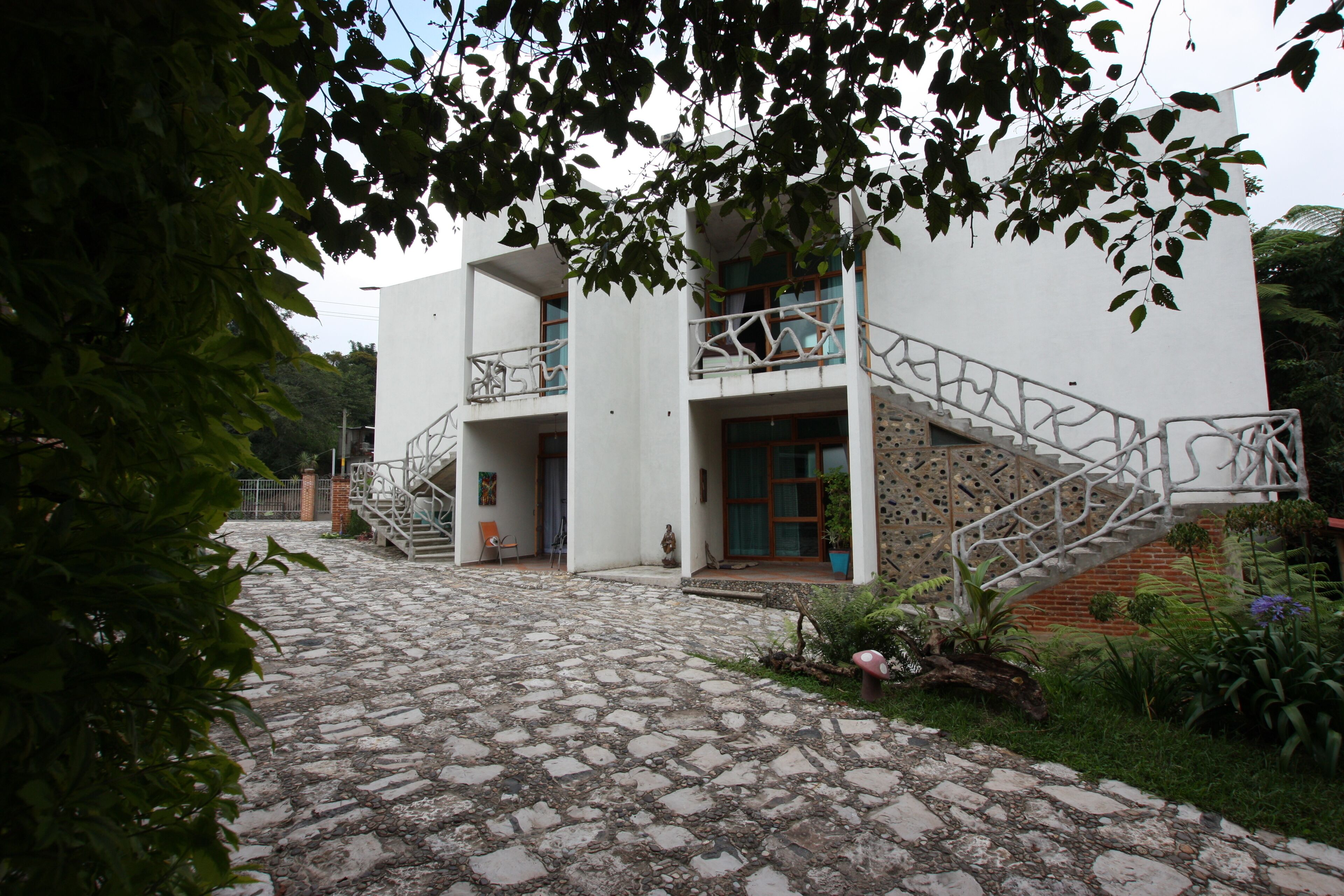 Un edificio moderno de dos plantas con un patio empedrado de piedra, un balcón con baranda blanca y una escalera que lleva al piso superior.