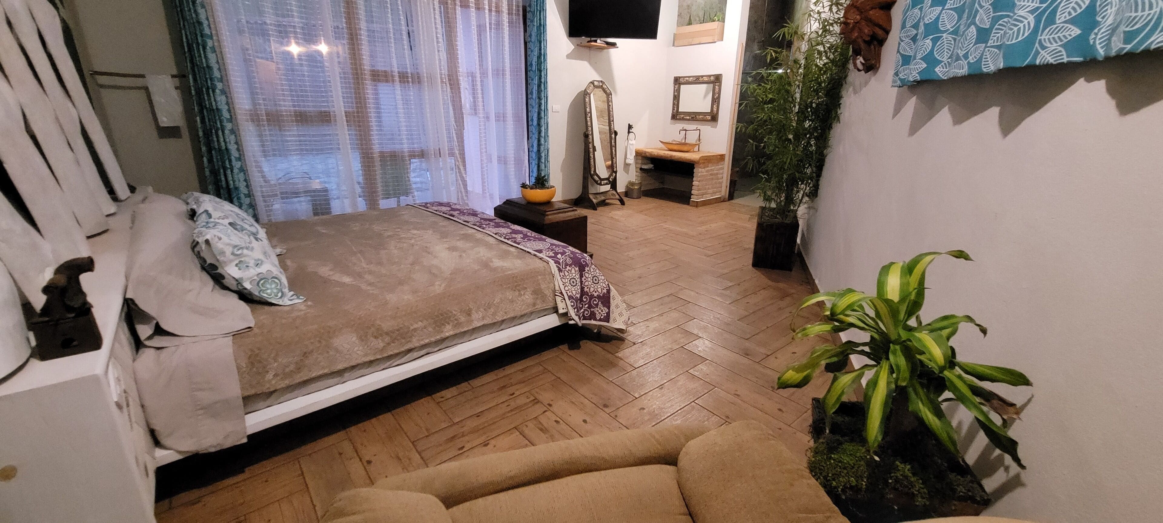 Un dormitorio con una cama, una mesita de noche, un espejo, una planta en maceta y suelo de madera.