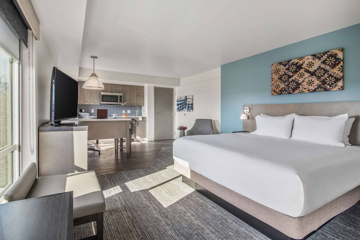 Een moderne hotelkamer met een groot bed, een bureau, een stoel, een televisie en een kitchenette op de achtergrond.