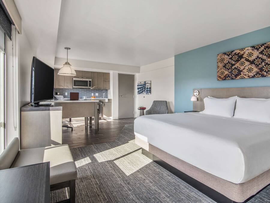 A modern hotel room with a large bed, a desk, a chair, a television, and a kitchenette in the background.