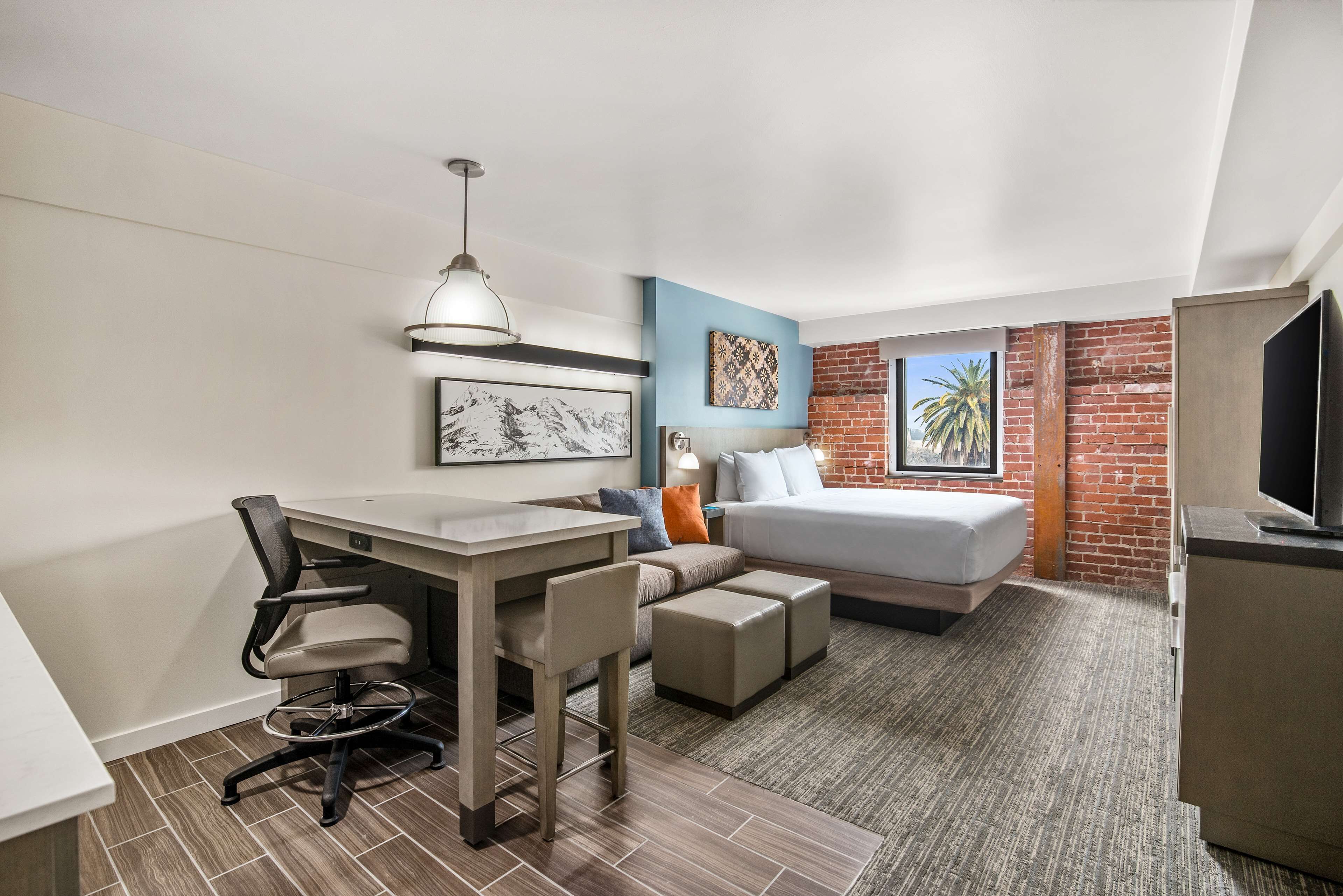 A compact hotel room with a bed, a desk, a chair, a television, and a small kitchenette.