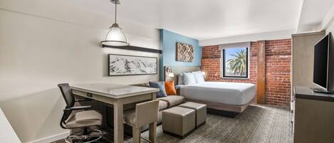 A compact hotel room with a bed, a desk, a chair, a television, and a small kitchenette.
