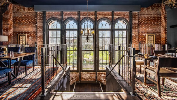 A spacious room with a brick wall, large arched windows, a chandelier, and a staircase leading to an outdoor area.