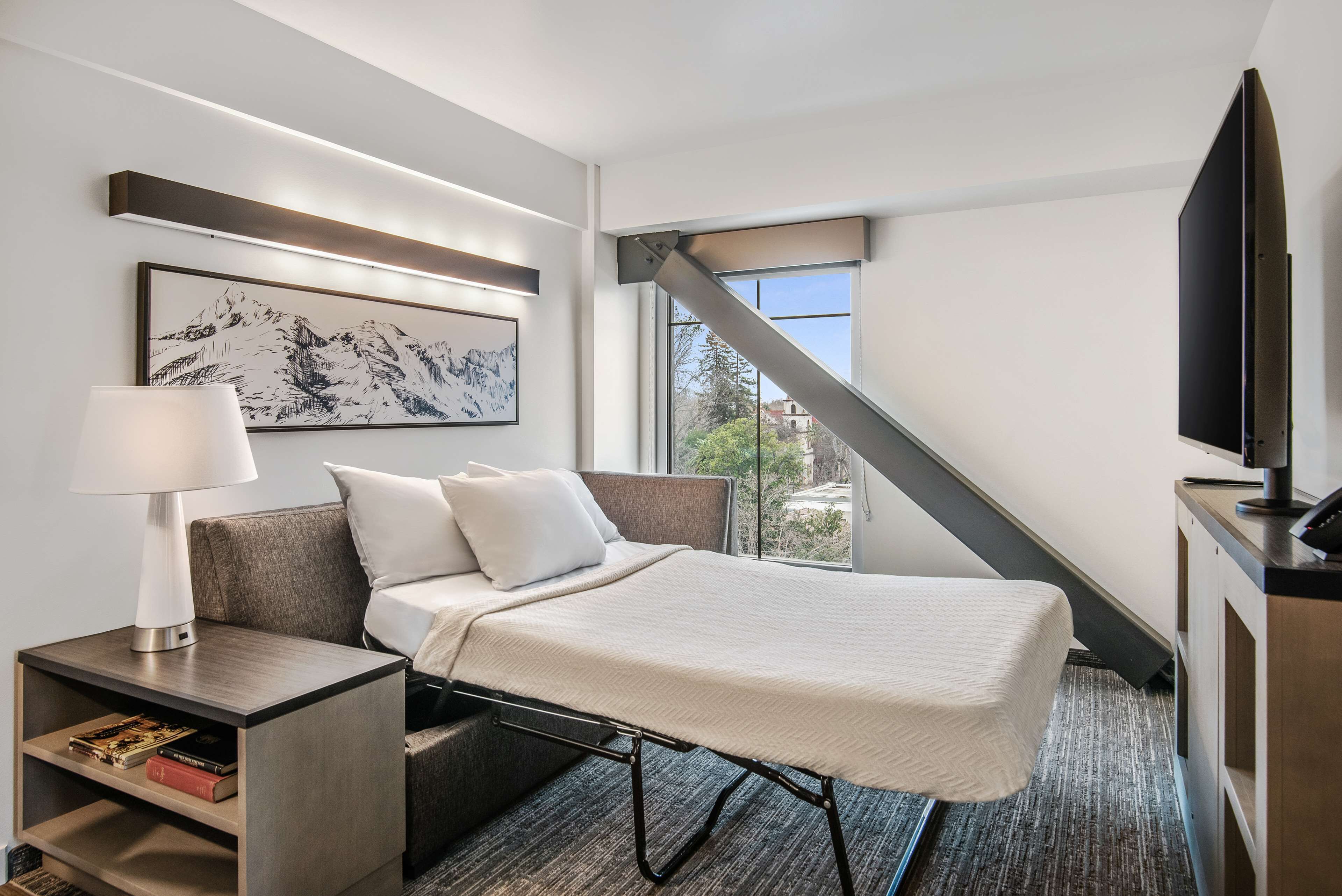 A modern hotel room with a bed, a nightstand with a lamp, a flat-screen TV, and a large window offering a view of the outdoors.