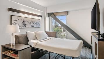 A modern hotel room with a bed, a nightstand with a lamp, a flat-screen TV, and a large window offering a view of the outdoors.