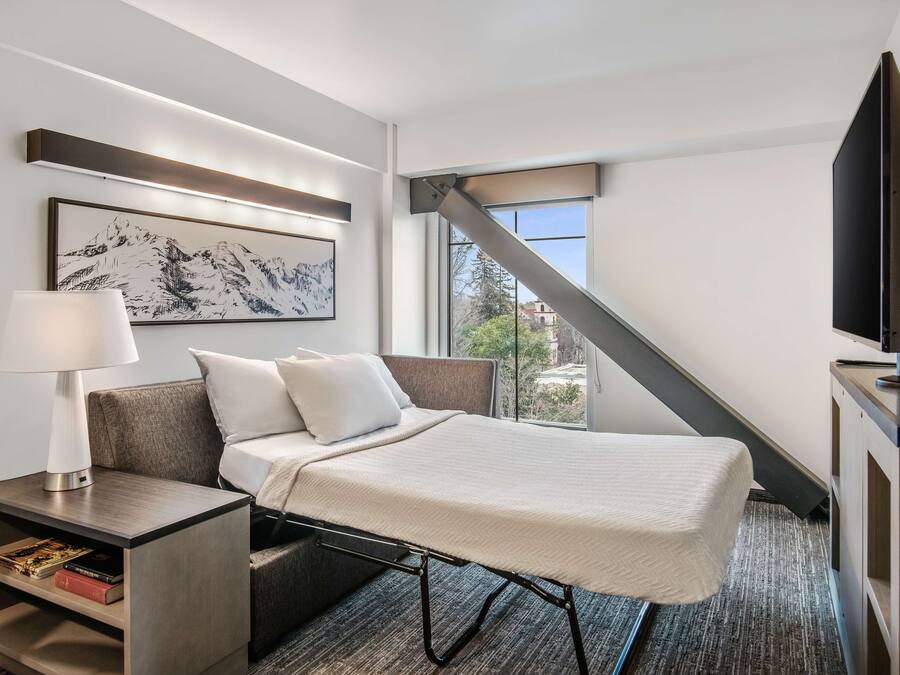 A modern hotel room with a bed, a nightstand with a lamp, a flat-screen TV, and a large window offering a view of the outdoors.