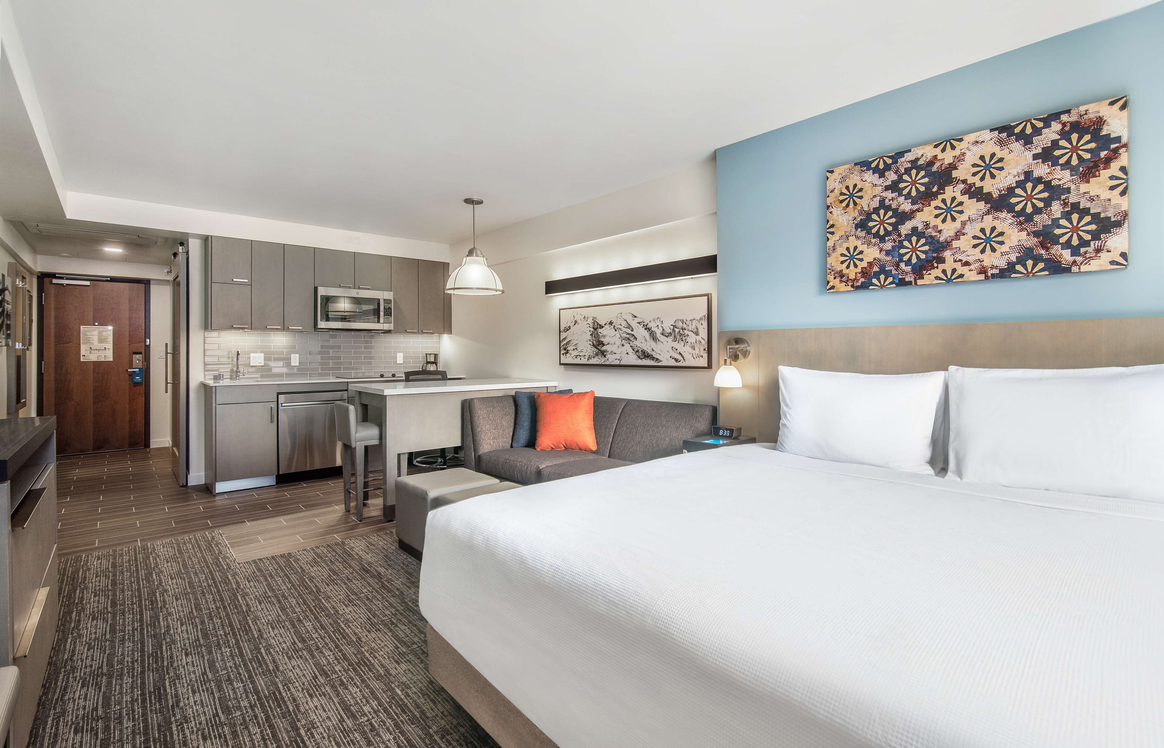A modern hotel room with a large bed, a kitchenette, a gray sofa, and a blue accent wall.