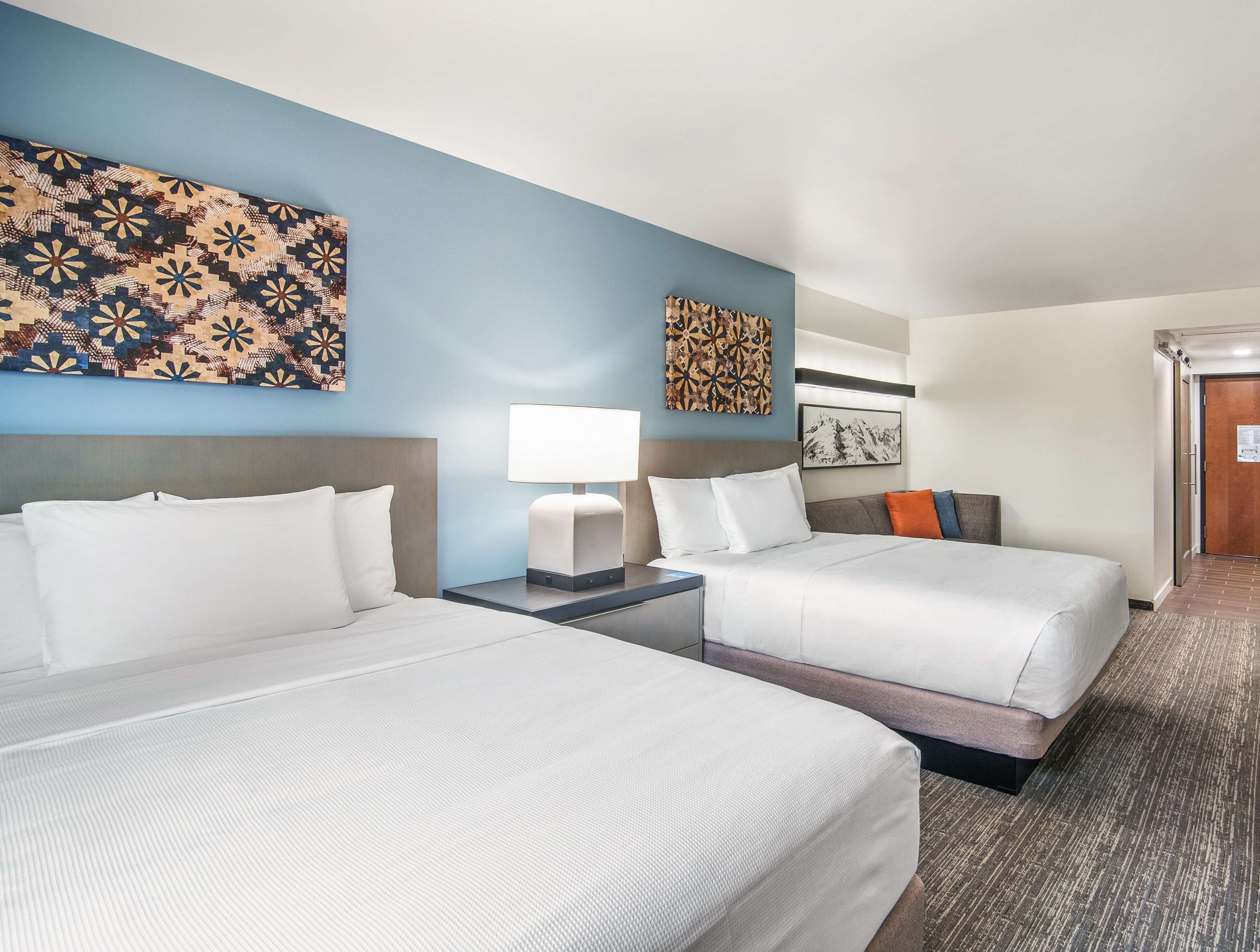A hotel room with two beds, a nightstand, a lamp, and artwork on the walls.