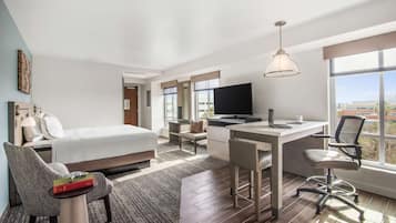 A modern hotel room with a bed, desk, and chair, a television, and large windows offering a city view.