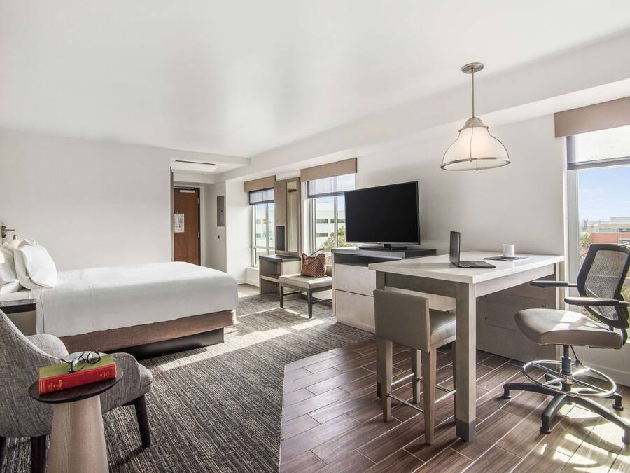 Une chambre d’hôtel moderne équipée d’un lit, d’un bureau et d’une chaise, d’une télévision et de grandes fenêtres donnant sur la ville.