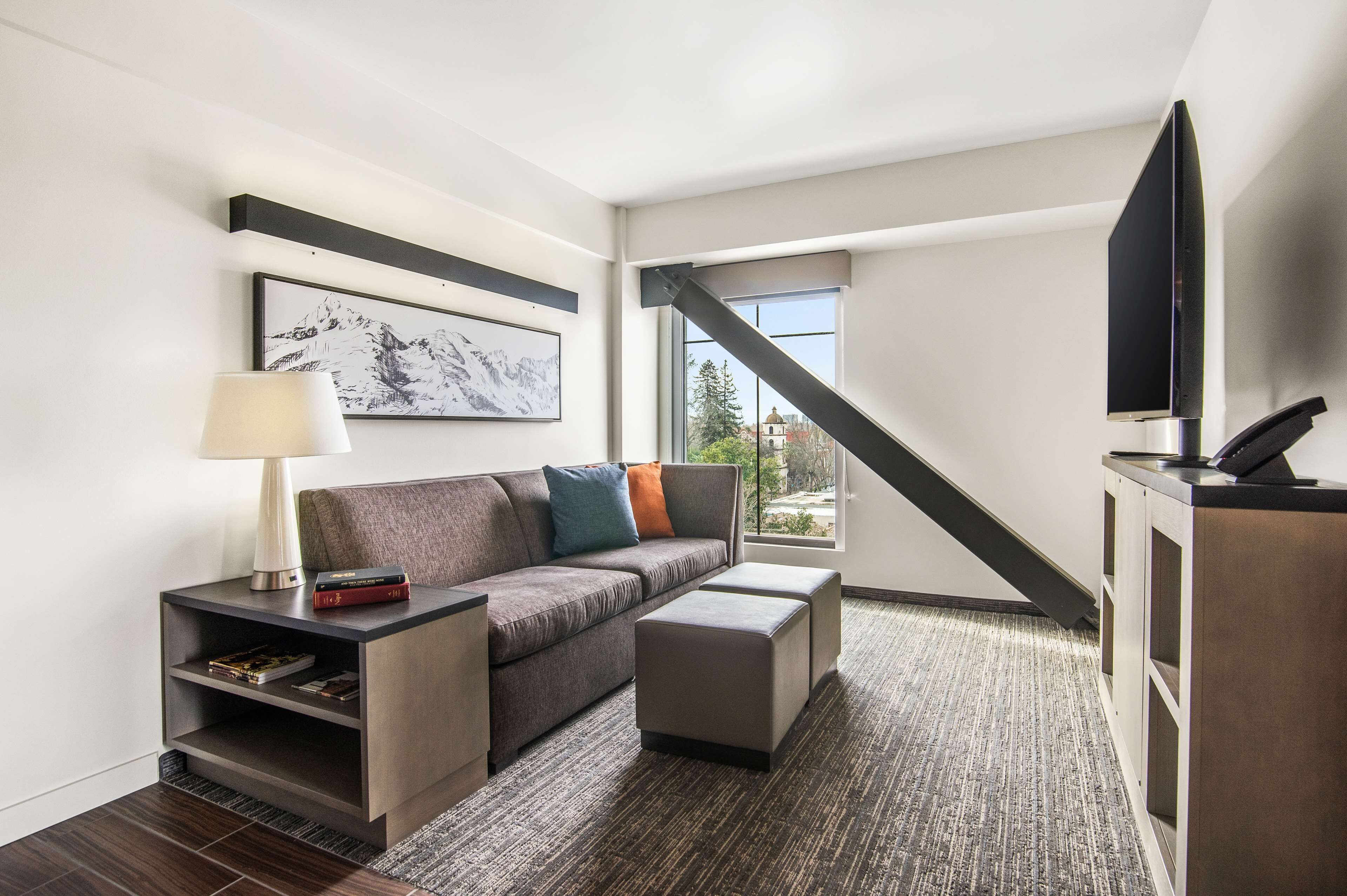 A modern hotel room with a sofa, a small table with a lamp, a flat-screen TV, and a large window offering a view of the outdoors.
