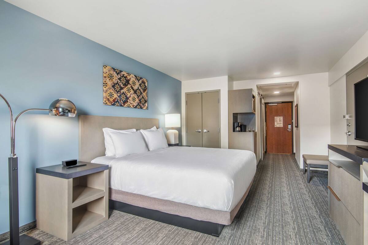 Een hotelkamer met een bed, nachtkastjes, een bureau, een televisie en een deur naar een gang.