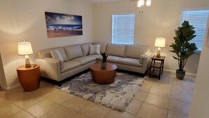 Una sala de estar con un sofá seccional color beige, dos mesas auxiliares con lámparas, una mesa de centro, una alfombra mullida, una planta en maceta y un cuadro de una escena de playa.