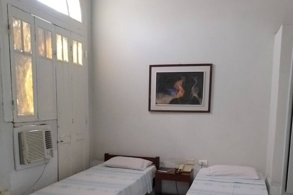 A small, clean room with two single beds, a chair, a small table, and a window with a view.