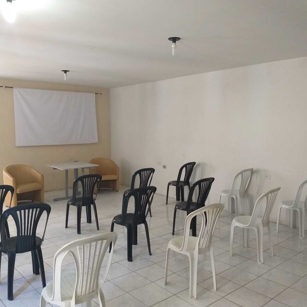 Una habitació amb un paviment de rajoles blanques, diverses cadires de plàstic, una taula petita i un pancartà blanc a la paret.