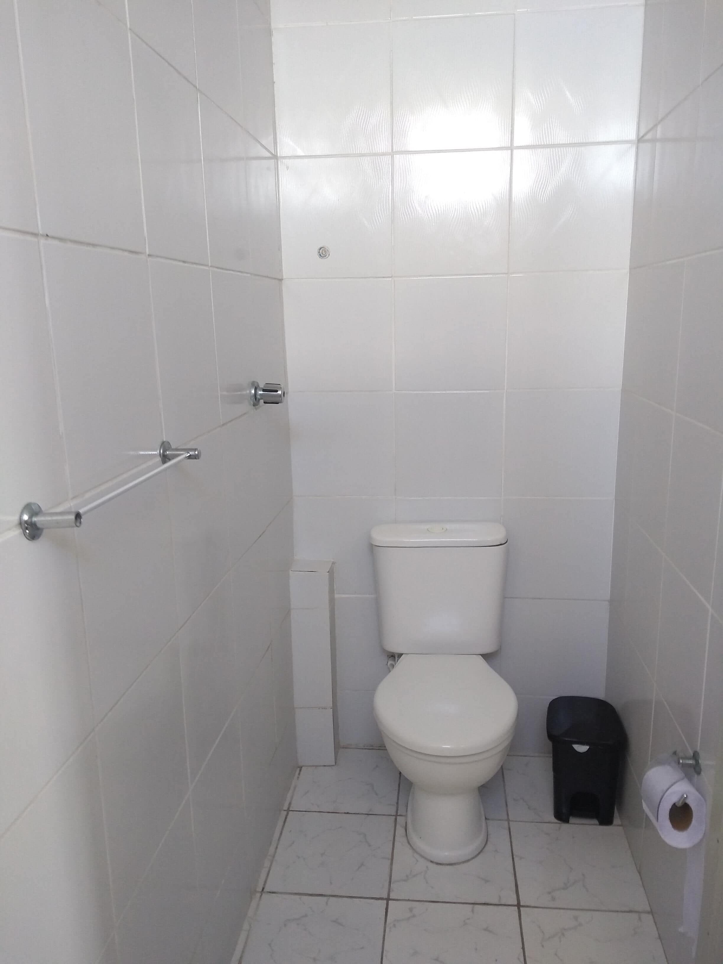 A small, white-tiled bathroom with a toilet, a trash bin, and a toilet paper holder.