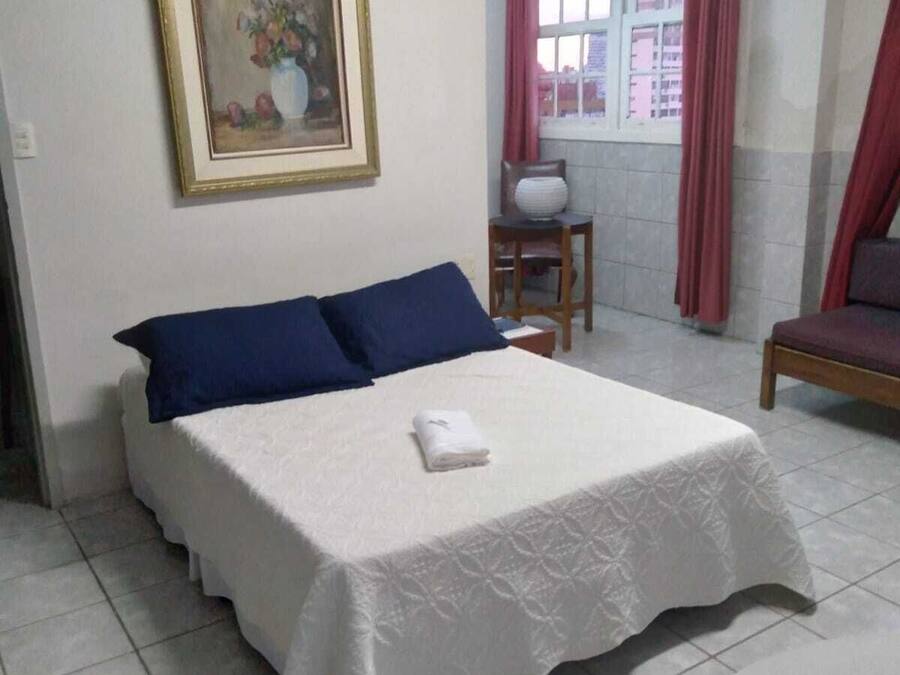 A hotel room with a bed, a chair, a table, a window with curtains, and a painting on the wall.