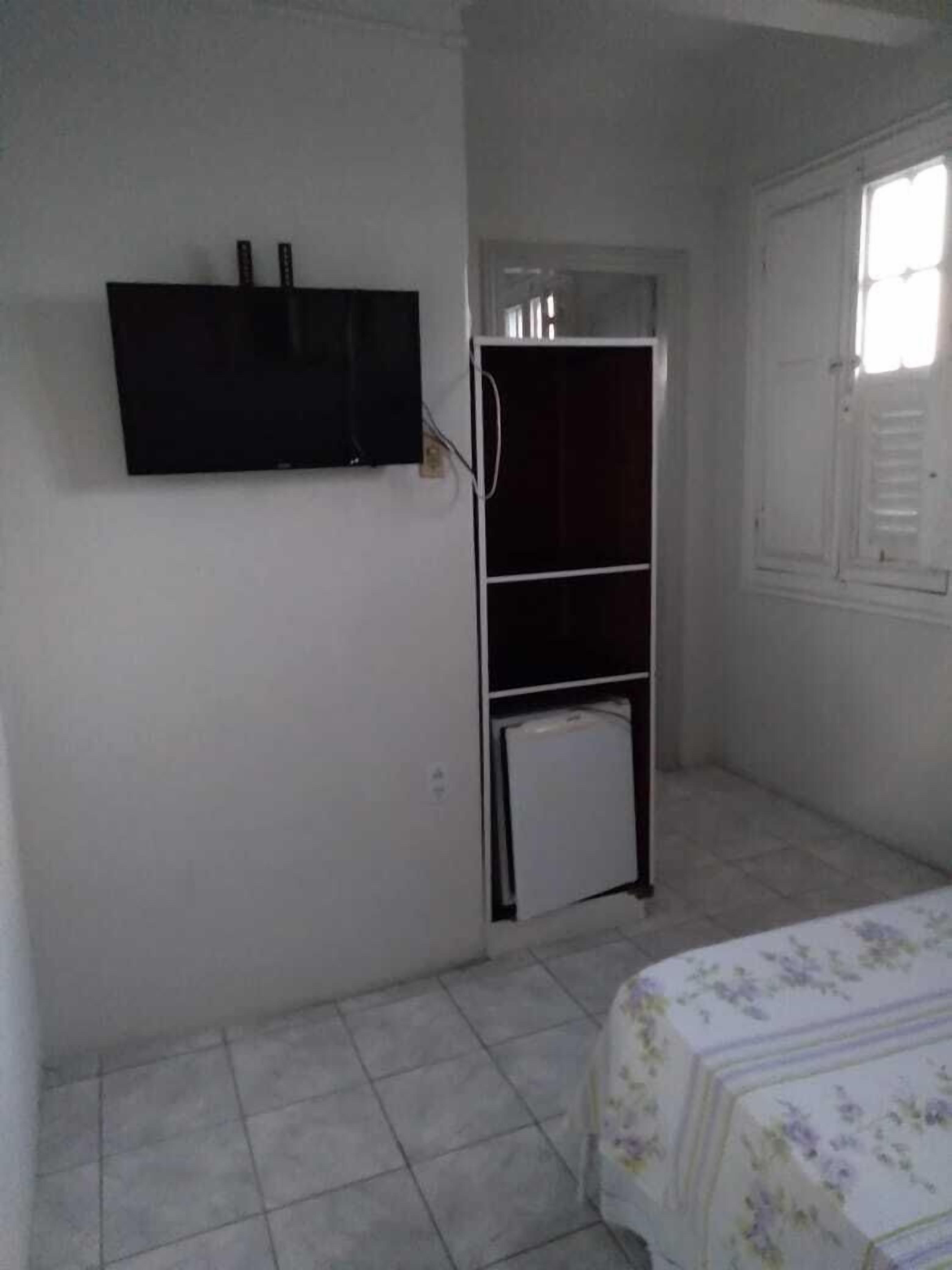 Una habitación pequeña y escasamente amoblada con una cama, un televisor en la pared y un refrigerador.
