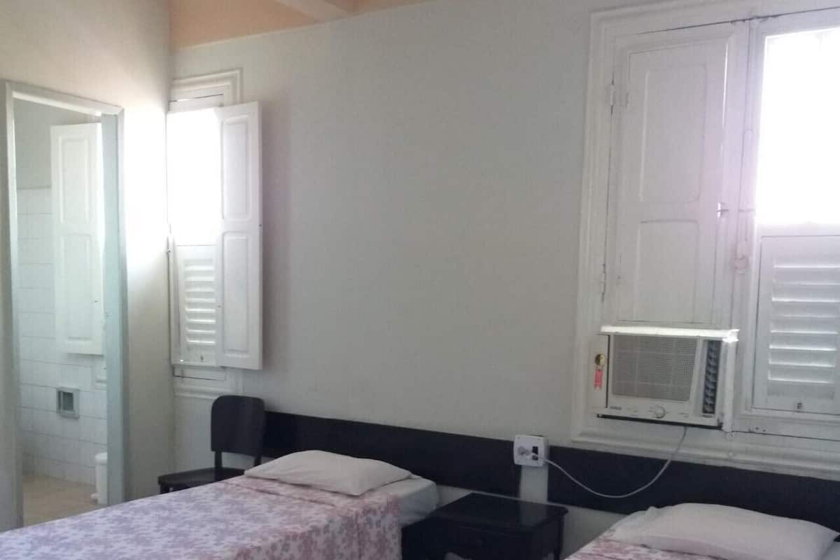 A room with two single beds, a small table, a chair, and a window with an air conditioning unit.