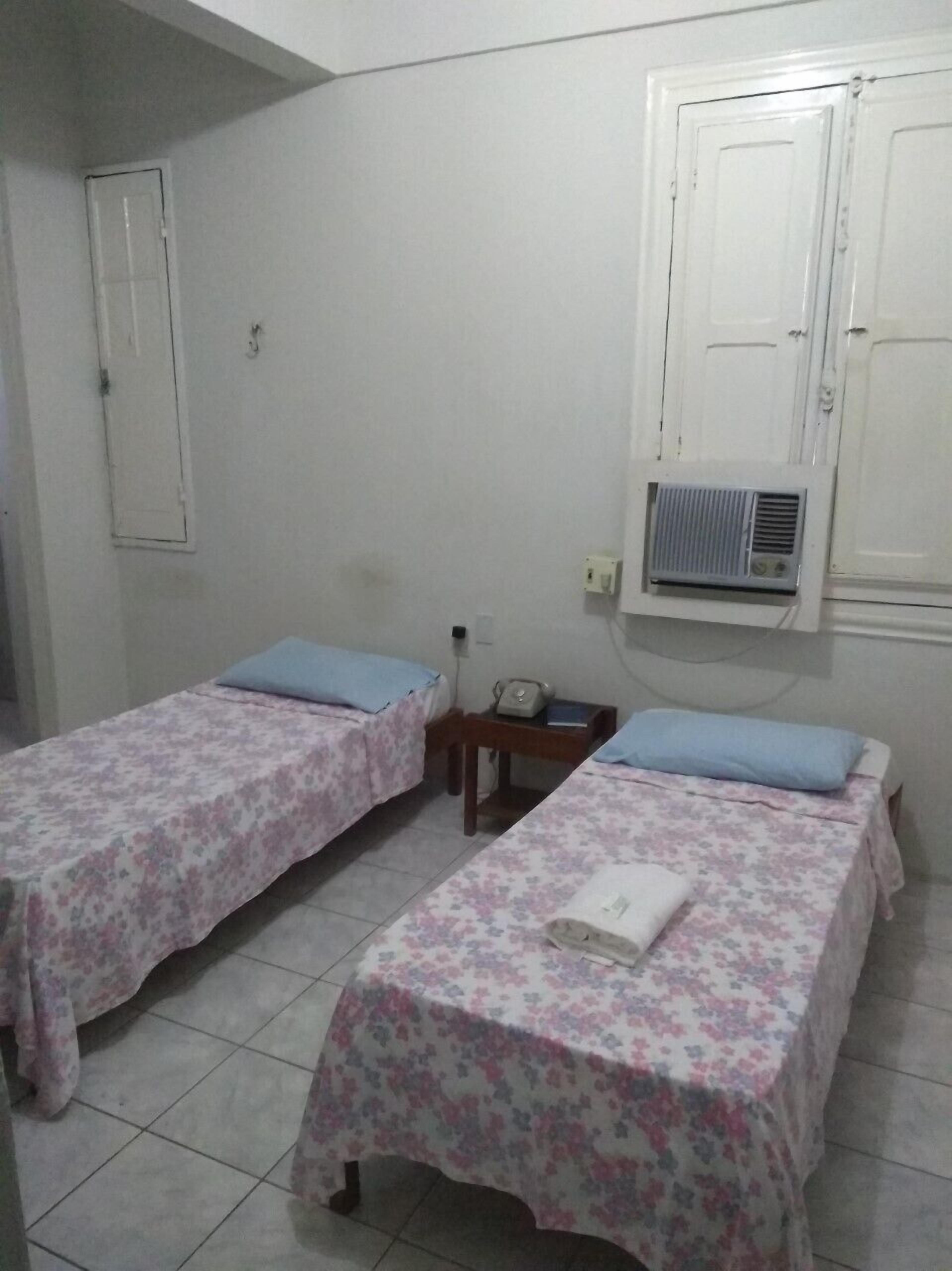 Una habitación pequeña y limpia con dos camas individuales, una mesita de noche con lámpara y aire acondicionado instalado en la pared.