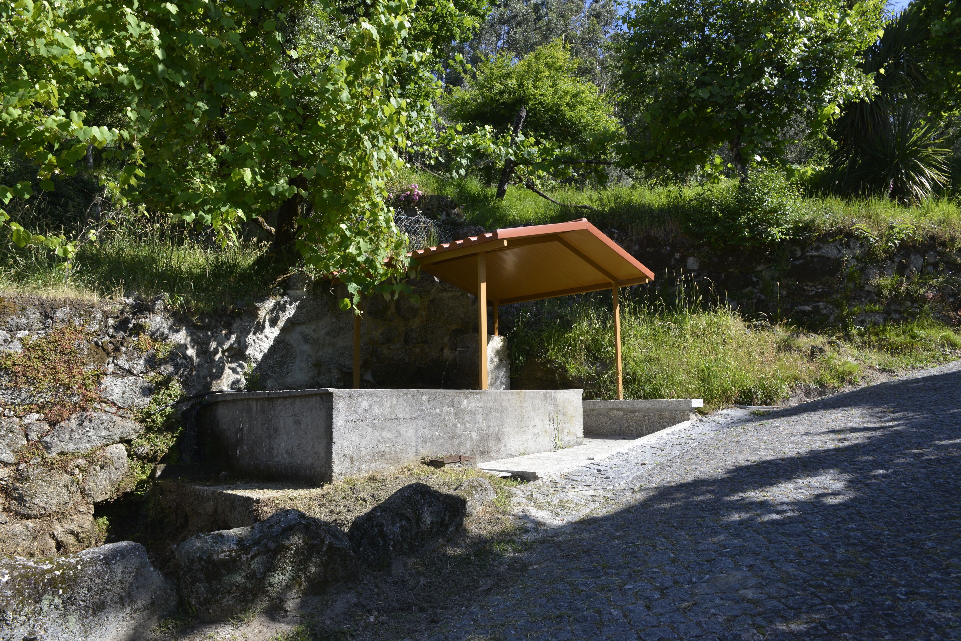A stone structure with a wooden shelter, surrounded by greenery and a cobblestone path.
