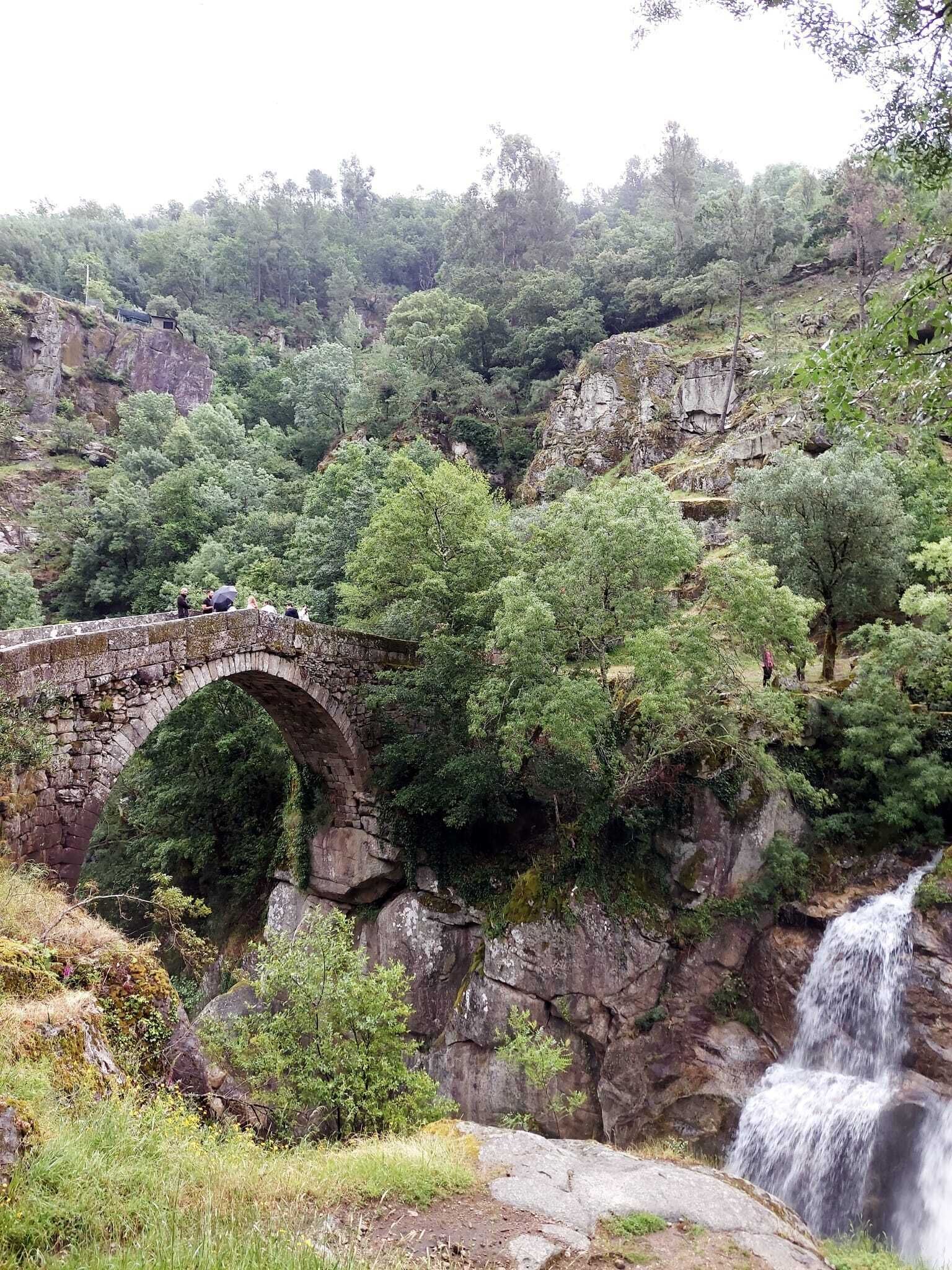Uma ponte de pedra sobre um rio, rodeada por vegetação exuberante e uma cascata.