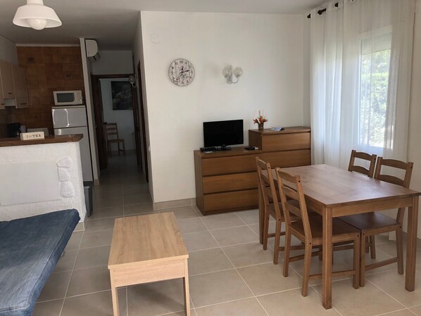 A compact living space with a kitchenette, a dining area with a wooden table and chairs, a small wooden side table, a TV on a wooden cabinet, and a wall clock.
