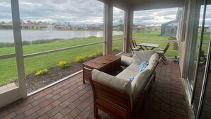 A covered patio with a sofa, chairs, and a table. It overlooks a body of water and a grassy area with houses in the background. - New Elegant Vacation Home in Fabulous Fort Myers (Fort Myers)