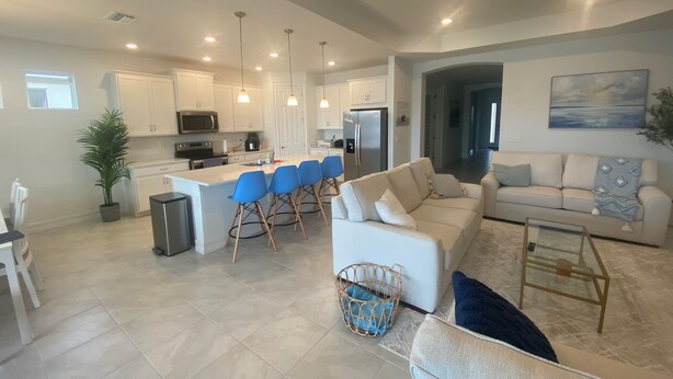 A spacious living area with a large white sofa, a coffee table, and a kitchen area with white cabinets and a central island with blue bar stools. - New Elegant Vacation Home in Fabulous Fort Myers (Fort Myers)