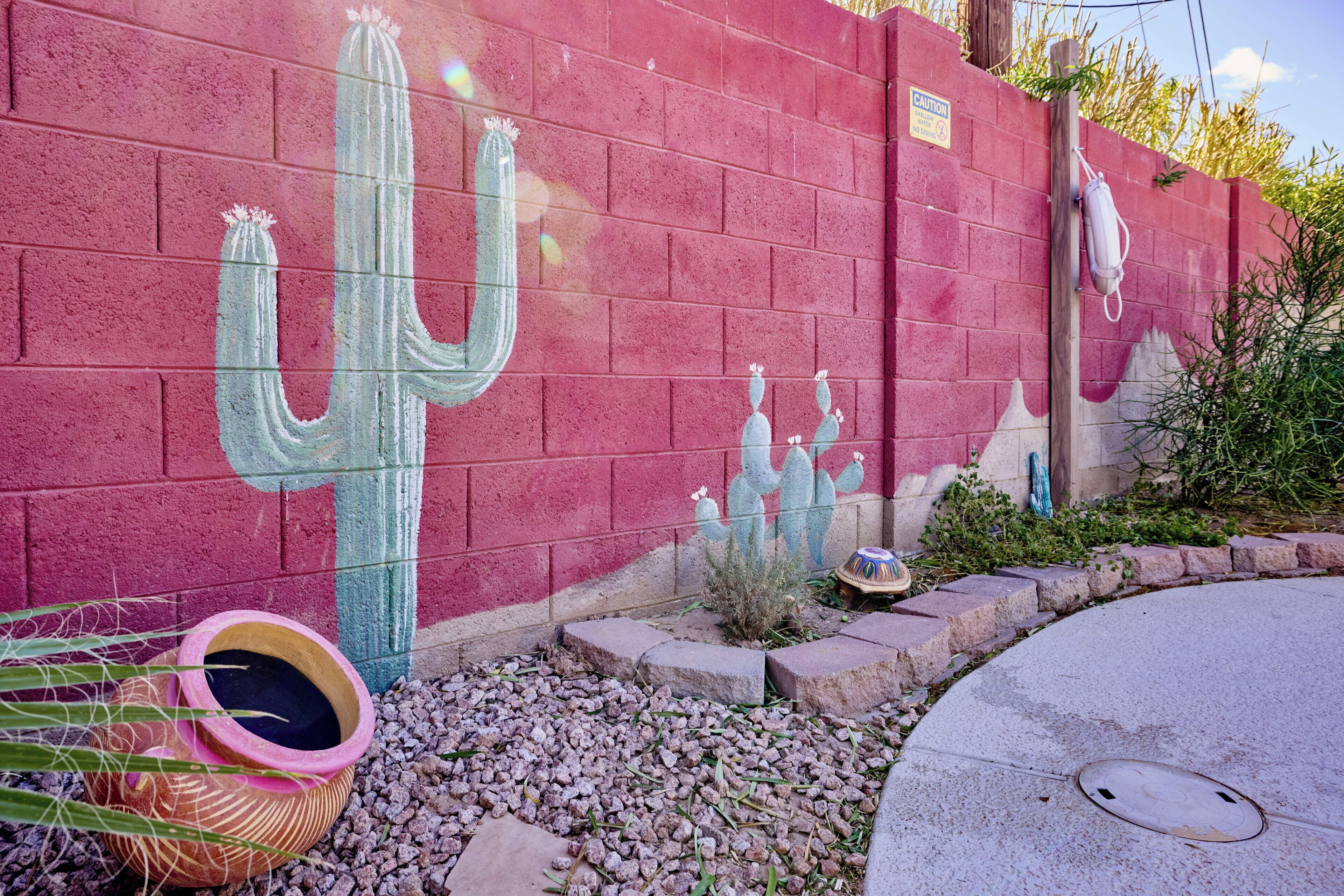 A red brick wall with a mural of cacti. There is a planter with a cactus and a small decorative item on the ground.