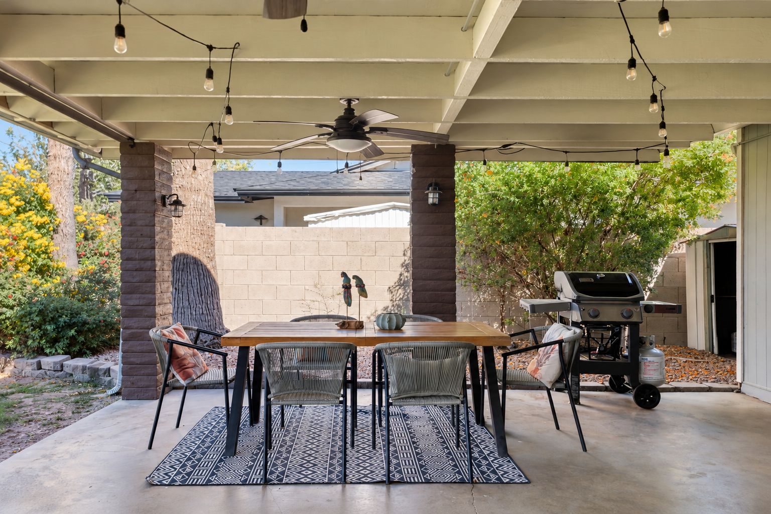 An outdoor patio with a table and chairs, a ceiling fan, string lights, and a barbecue grill.