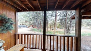 A wooden deck with a bench, a pine tree, and a view of a forest and parked vehicles. - Walk to Lifts | Wooded Area | Rustic Interior | French Henry B2 (Angel Fire)